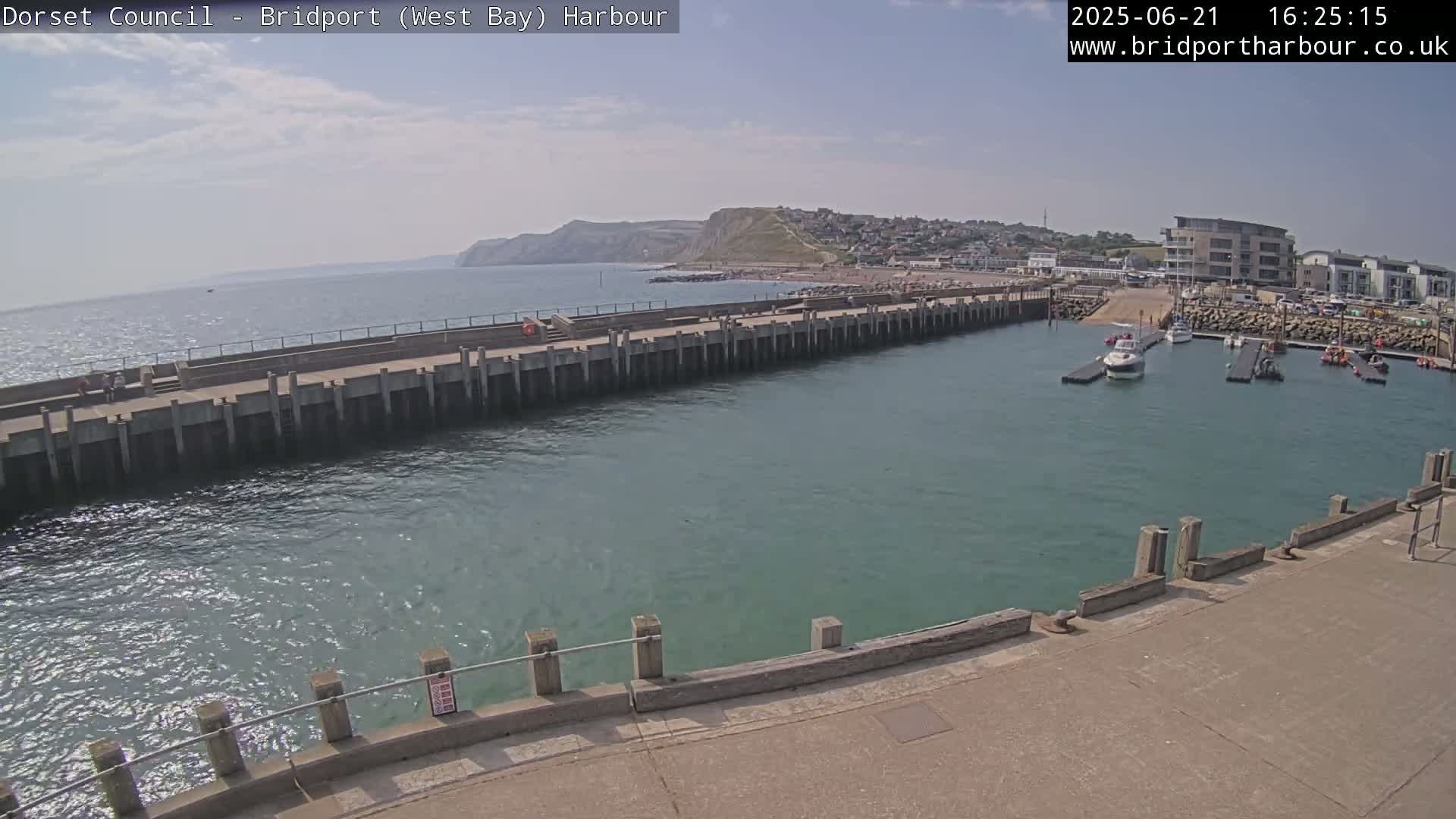 A harbor with calm, teal water is partially enclosed by a long concrete pier extending from a paved area, with a town and cliffs visible in the background under a mostly clear sky.