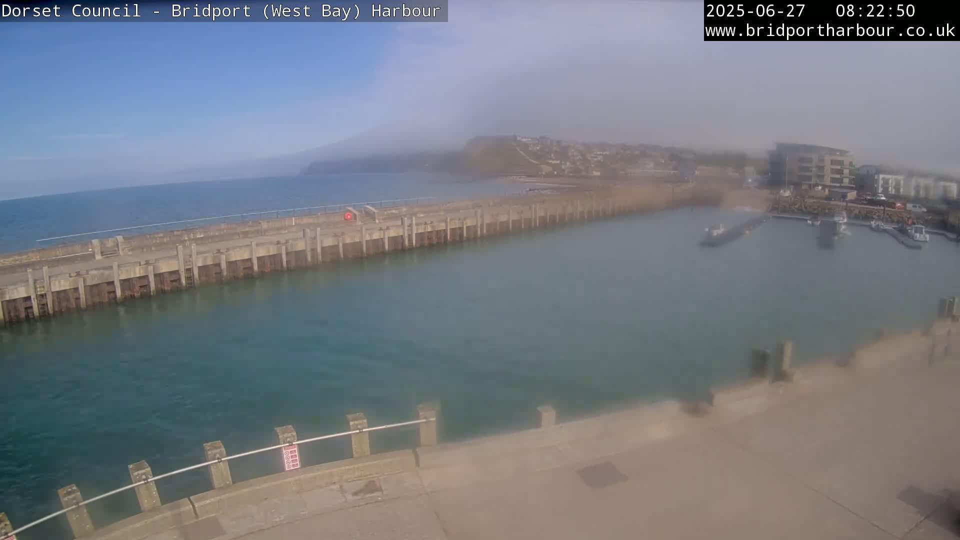 A hazy harbor scene shows a long concrete pier extending into calm, teal water, with a town and distant hills visible in the background.