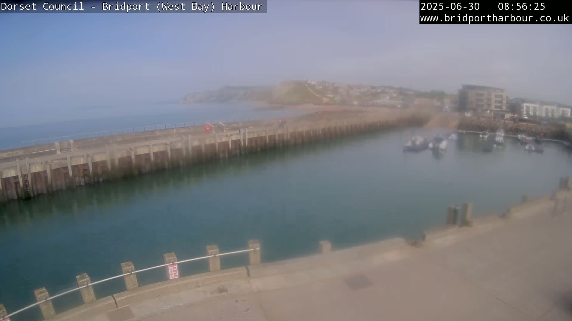 A hazy harbor with calm, teal water, a long concrete pier, and several boats moored in a marina, situated near a coastal town and hillside.