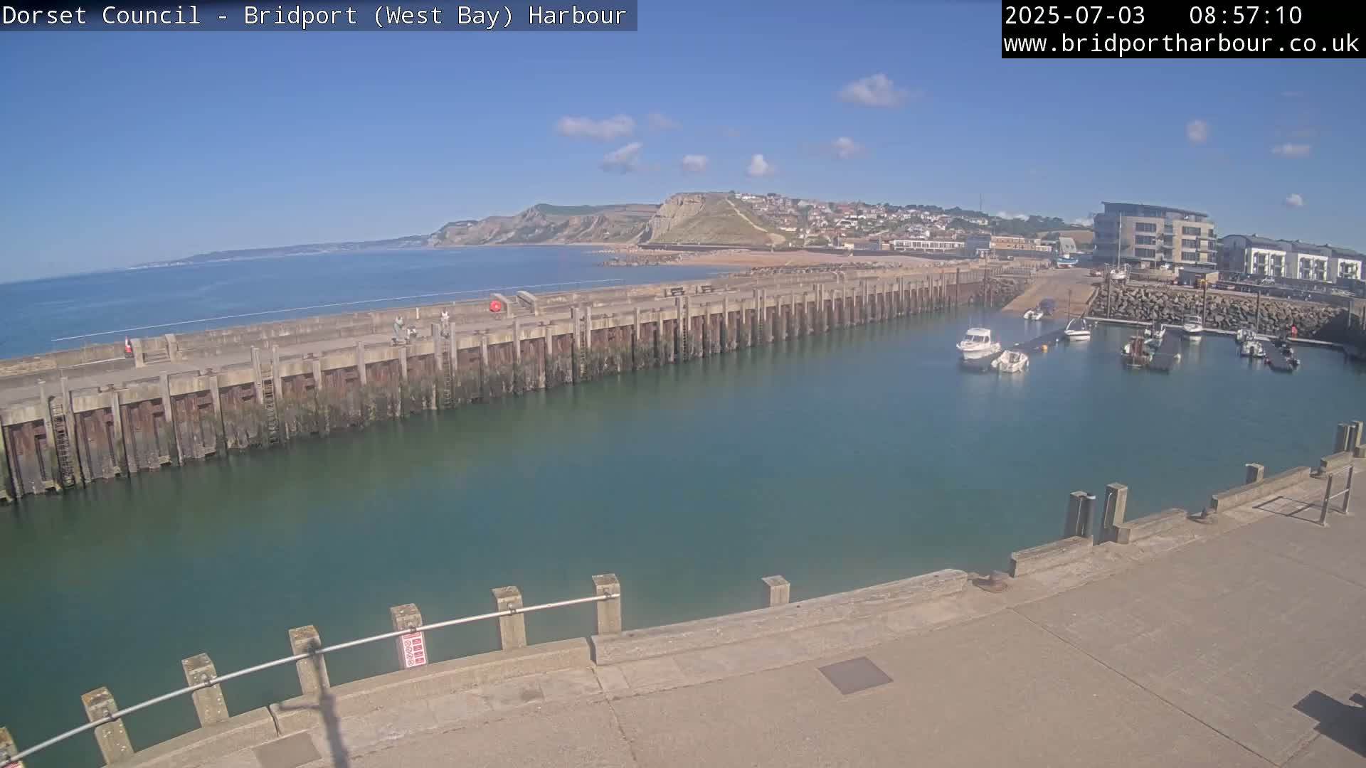 A harbor with several boats is shown on a sunny day, with a long wall separating the harbor from the ocean and a hilly coastline visible in the background.