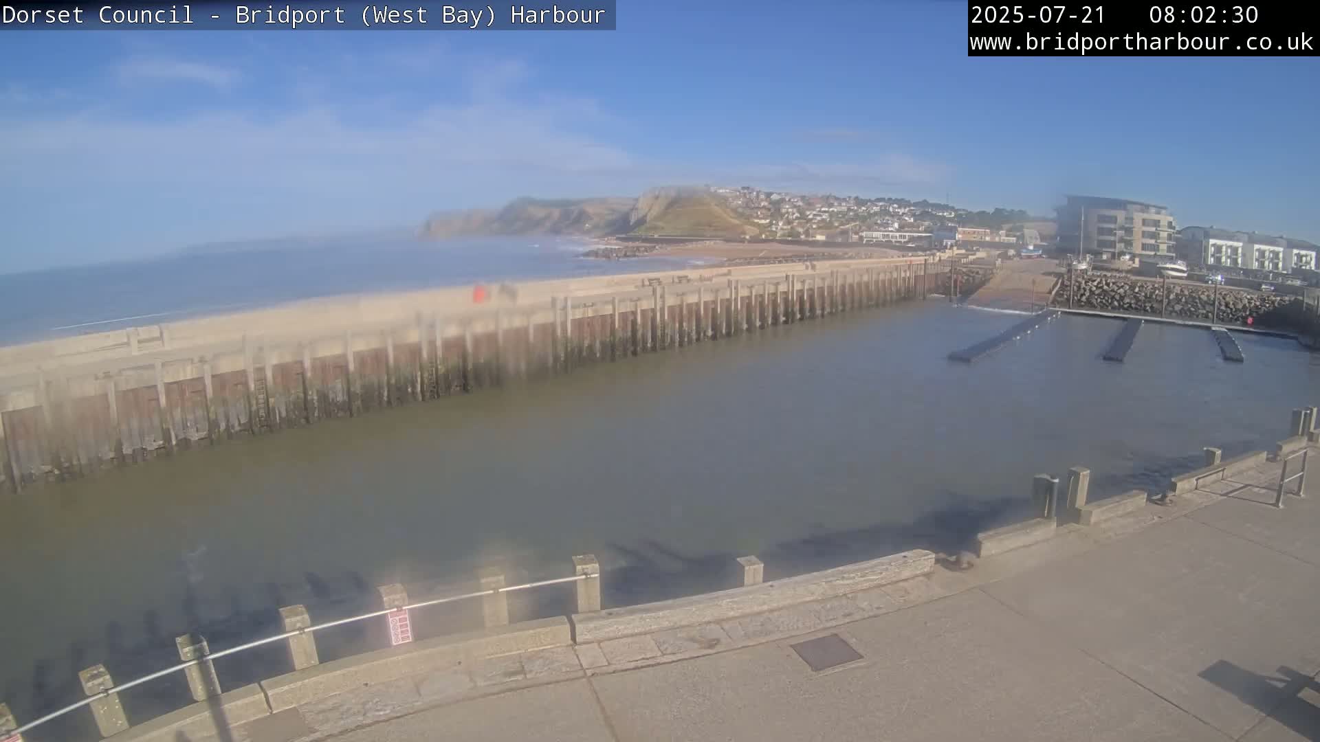 A harbor with calm water, a sandy beach, and a hill in the background under a mostly clear sky.