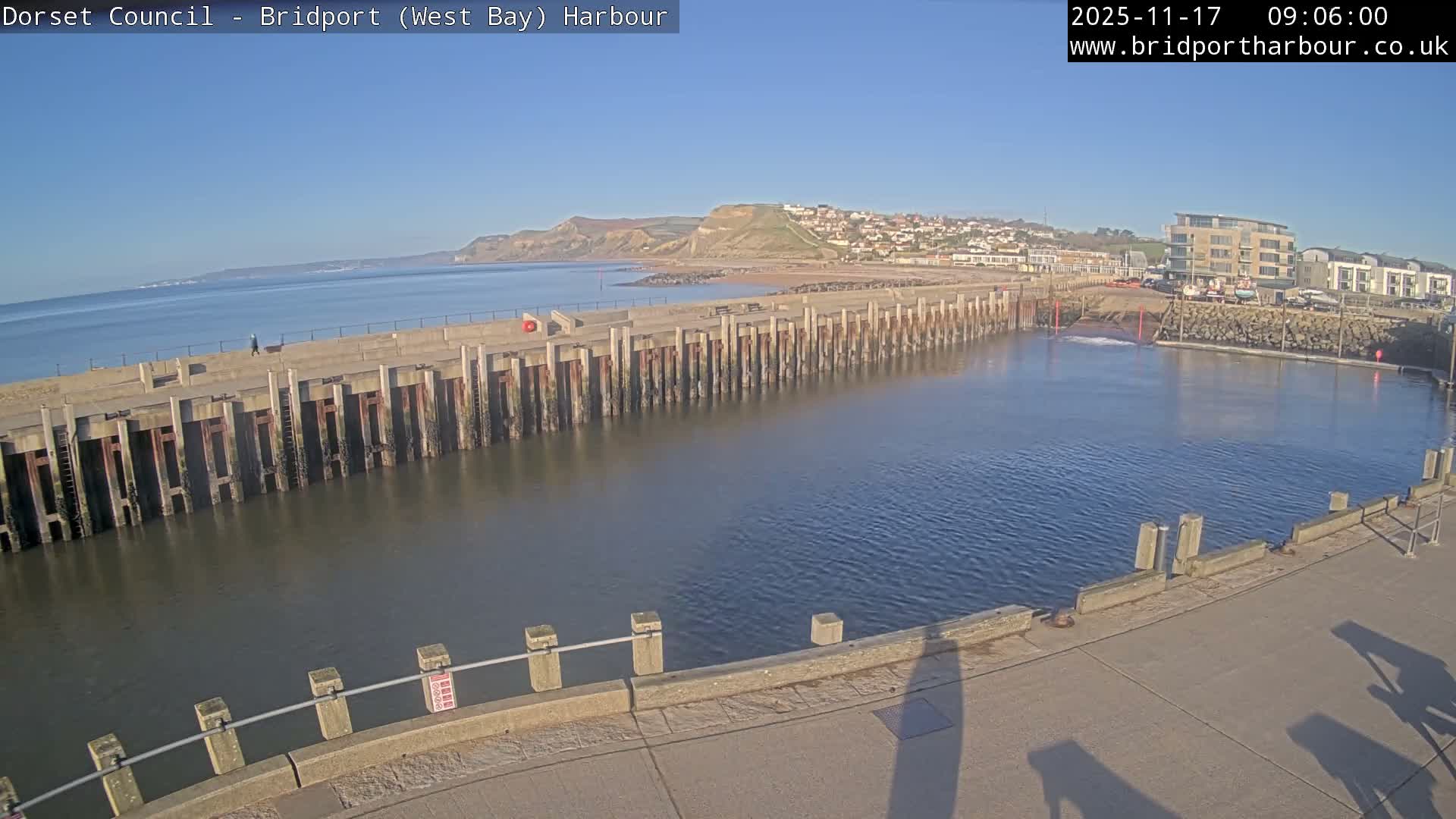 A sunny day illuminates a tranquil harbor with a long concrete pier, a distant town nestled on hills, and various buildings bordering the inner basin, all under a clear blue sky.