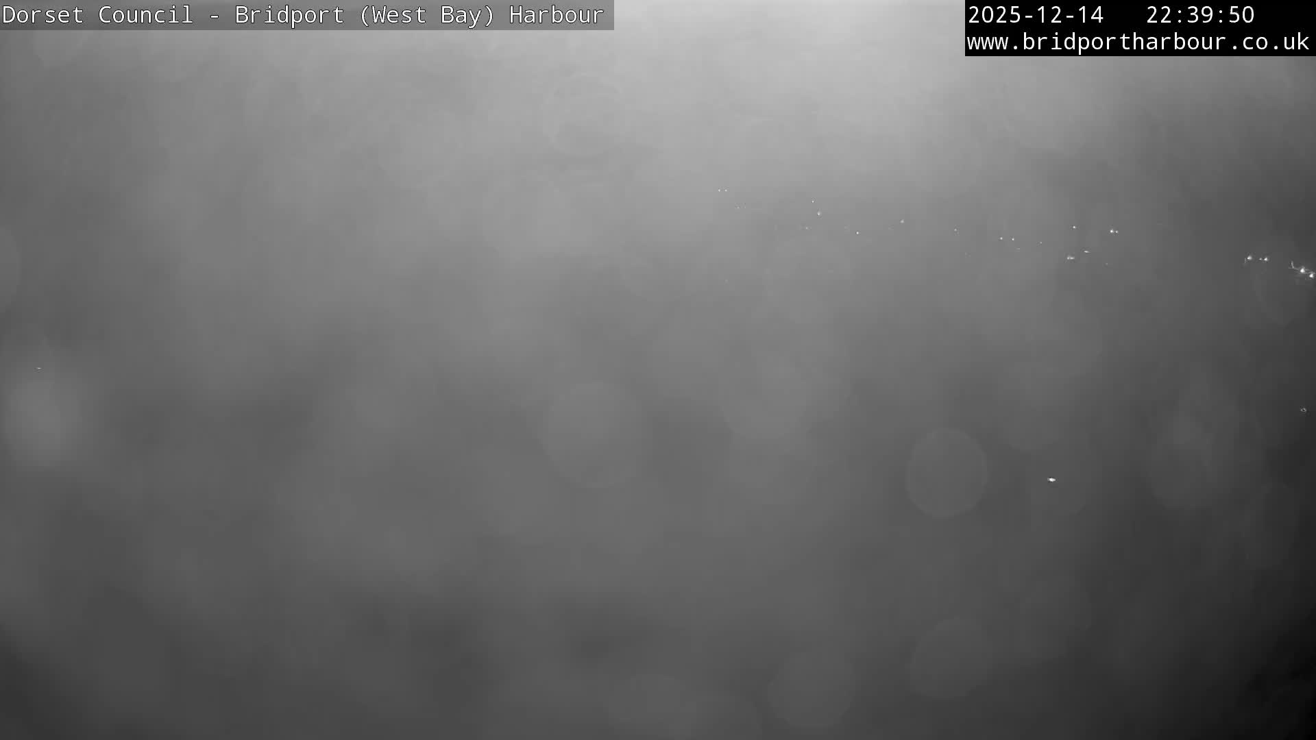 An overcast view of a coastal harbour features a long pier extending into choppy grey water, with a distant town built into hills and cliffs, and modern buildings lining the right side of the frame.