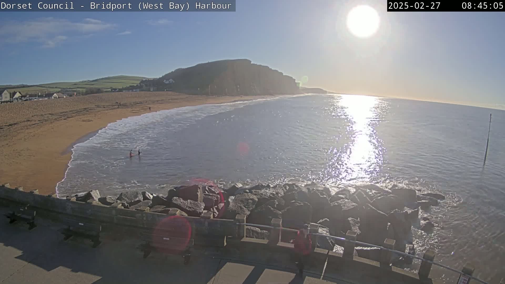 A sunny day at a beach with calm waves lapping a sandy shore, backed by a cliff face and a rocky breakwater.