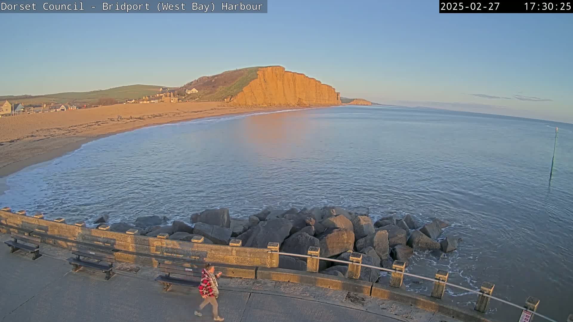 A sandy beach with calm, gently lapping waves, a person walking along a sea wall, and a large, reddish cliff in the background under a clear, twilight sky.