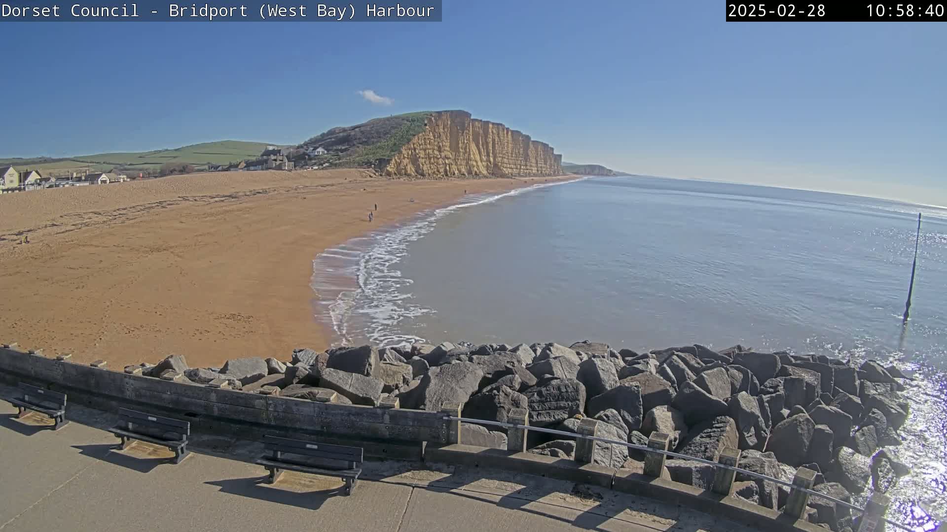 A sandy beach stretches out to a calm sea under a clear blue sky, backed by a dramatic cliff face.