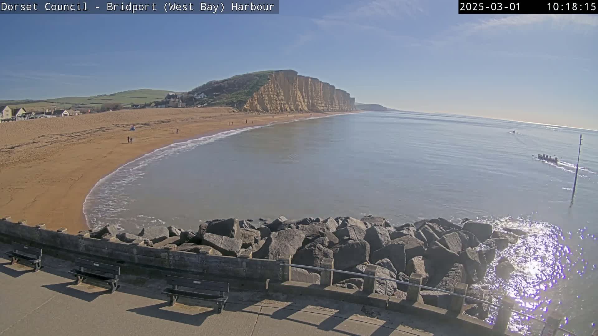 A sandy beach curves gently around a rocky breakwater, meeting a calm sea under a clear blue sky, with high cliffs visible in the distance.