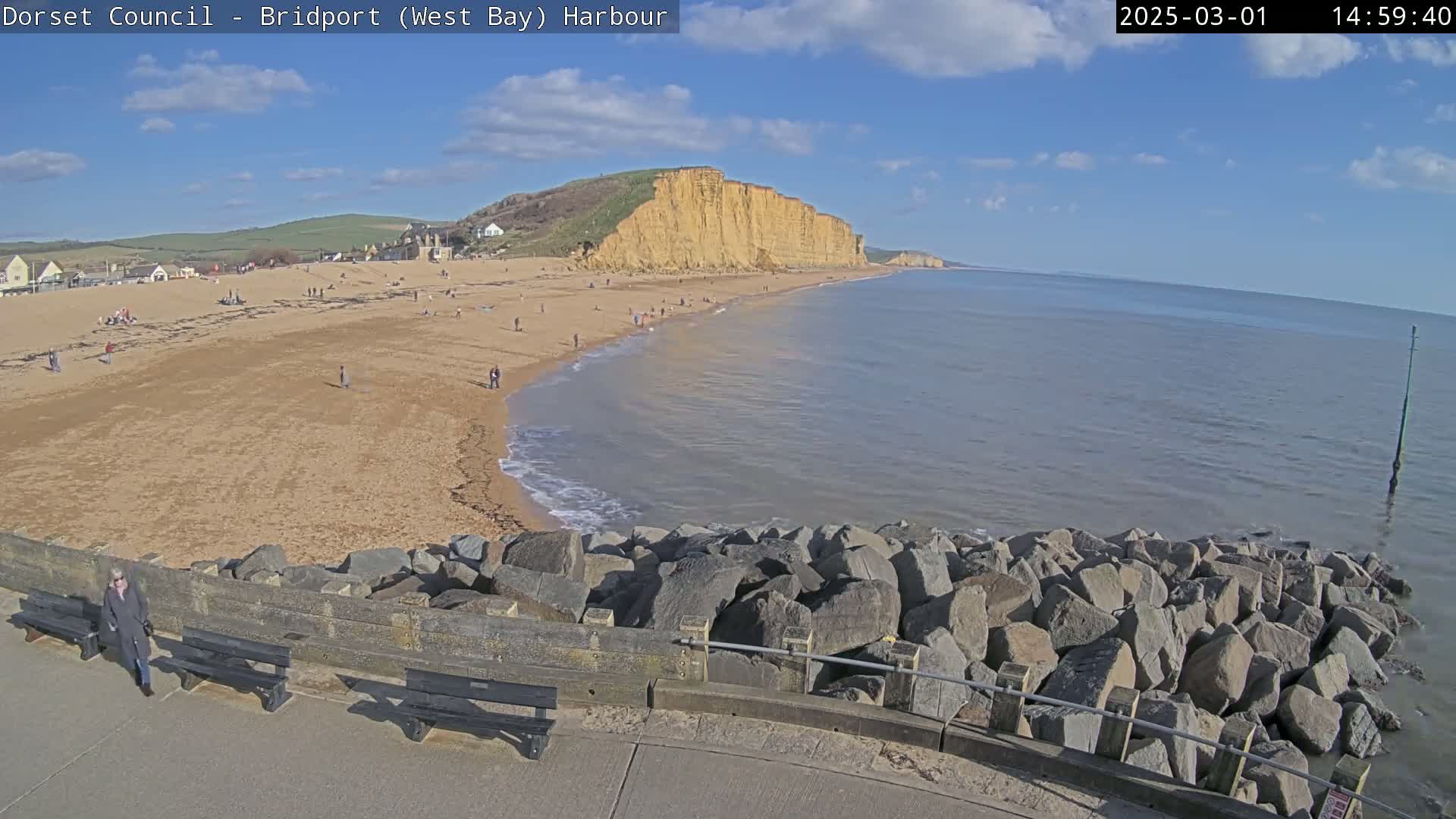 A sandy beach with people on it stretches beneath a large cliff face next to a calm sea under a mostly sunny sky.
