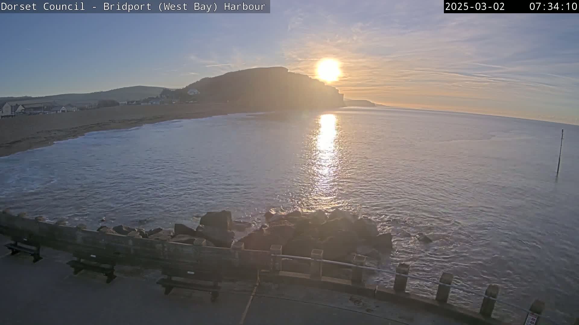 A sunrise over a calm bay, showing a sandy beach, cliffs, and a harbor wall with benches.