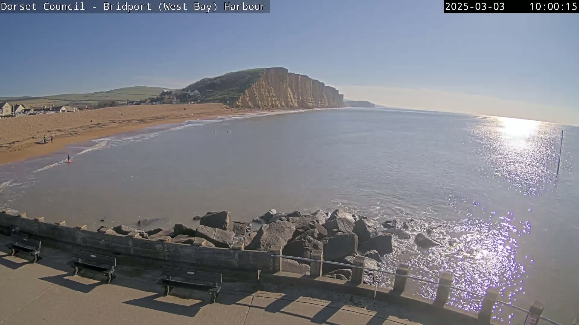 A sunny day at a harbor shows a sandy beach, calm water, and large cliffs in the distance.