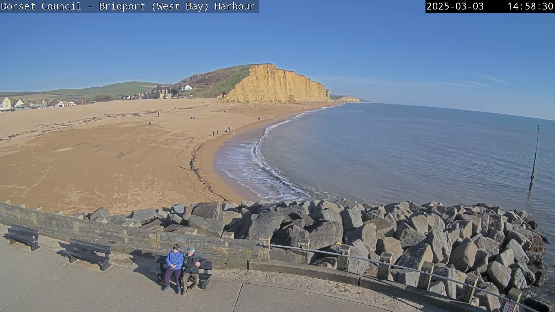 A sunny day at a beach with a sandy shore, gently lapping waves, and large cliffs in the background; two people sit on a bench overlooking the scene.