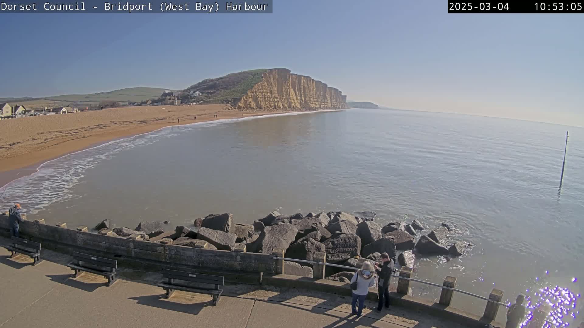 A sandy beach with calm, light-grey water meets a high cliff face under a clear, sunny sky, with a few people visible on the beach and near a low stone wall.