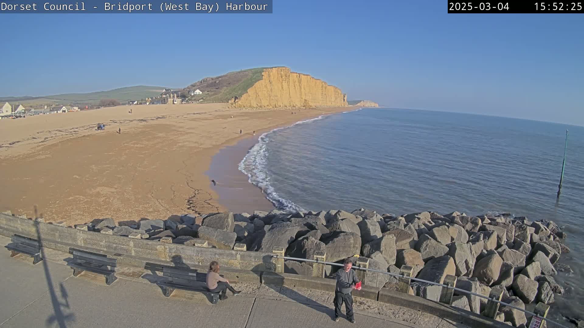 A sandy beach stretches beneath a sunny sky, meeting a calm sea that gently laps against a rocky breakwater where two people stand near empty benches.