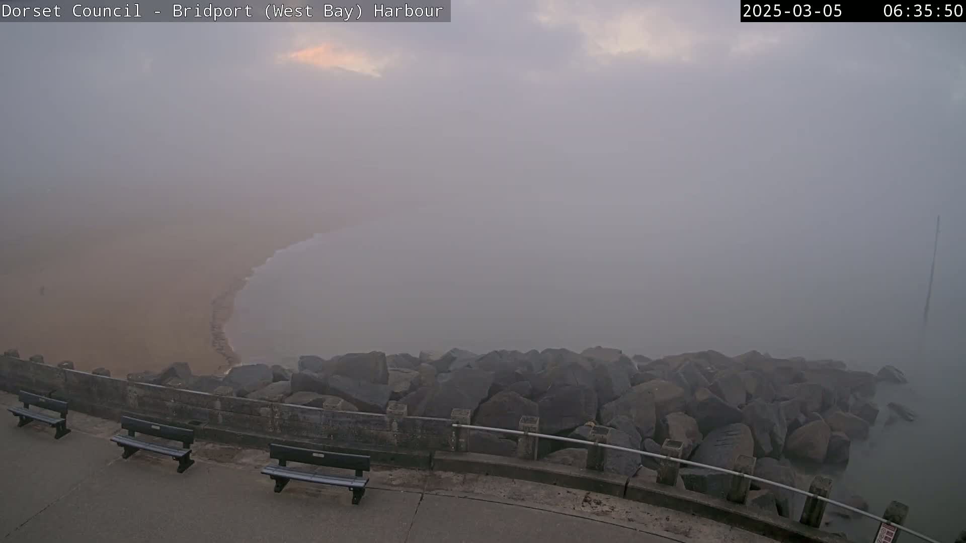 A foggy harbor scene shows a sandy beach curving into the water, partially obscured by a thick fog, with several benches and a rock barrier along a paved walkway.
