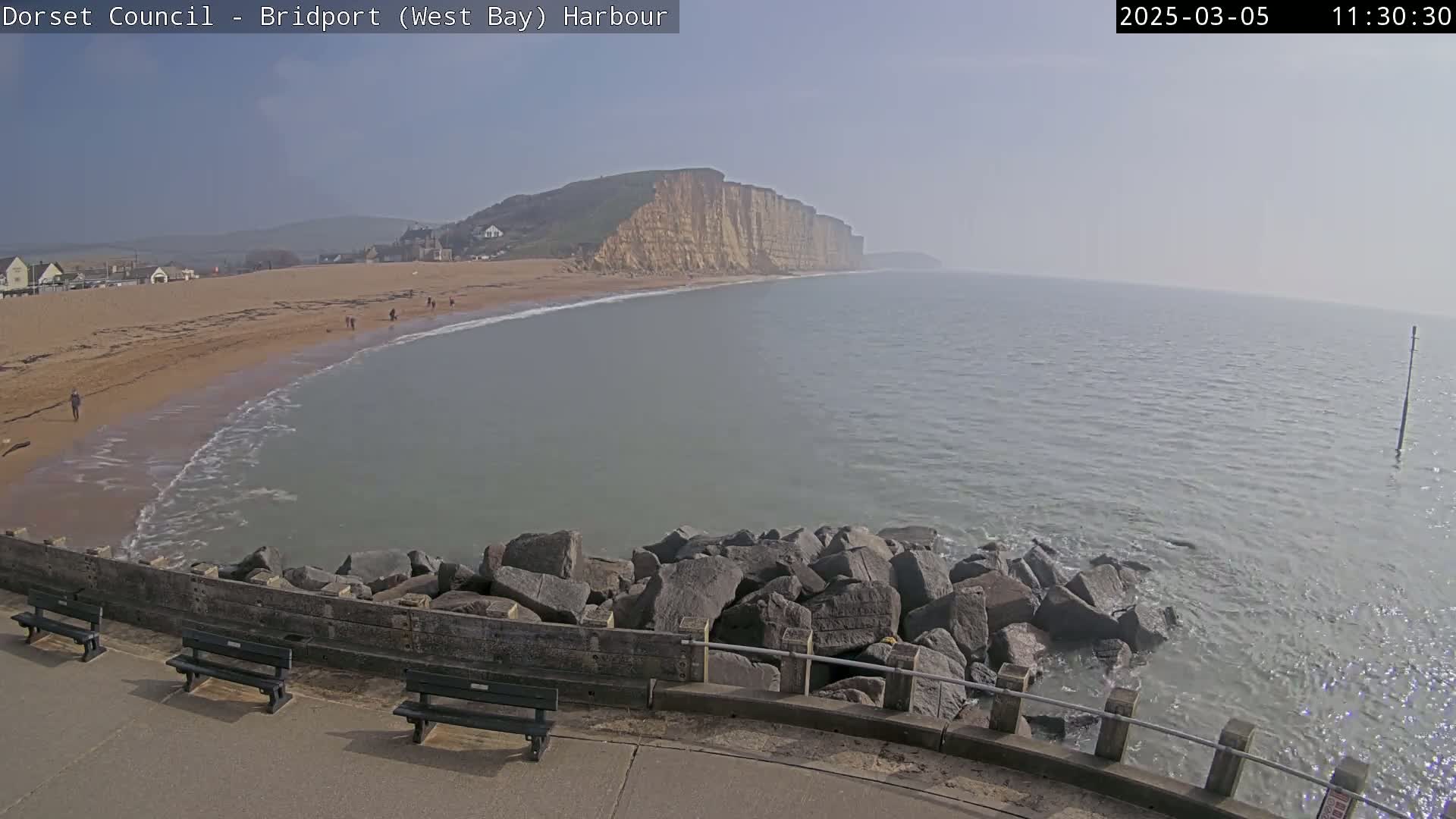 A hazy, sunny day reveals a beach with a few people, a calm sea with small waves lapping against a rocky breakwater, and tall cliffs in the distance.