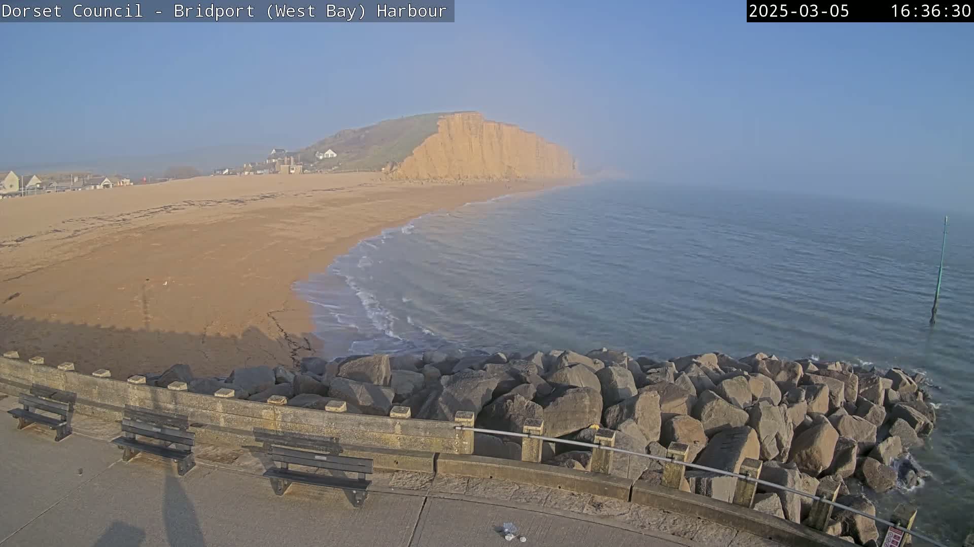 A sandy beach meets a calm, hazy sea, backed by a large cliff face partially obscured by fog, with a rocky seawall and benches in the foreground.