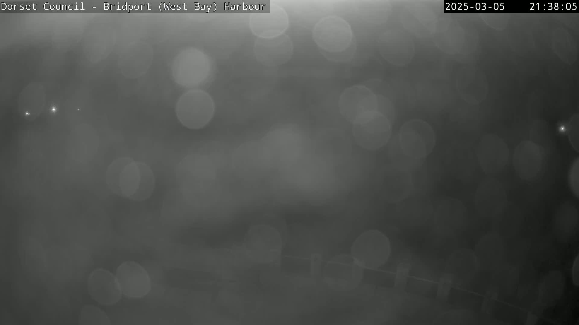 A blurry, dark gray image shows out-of-focus lights and a suggestion of a dark, possibly wet surface under a nighttime sky.