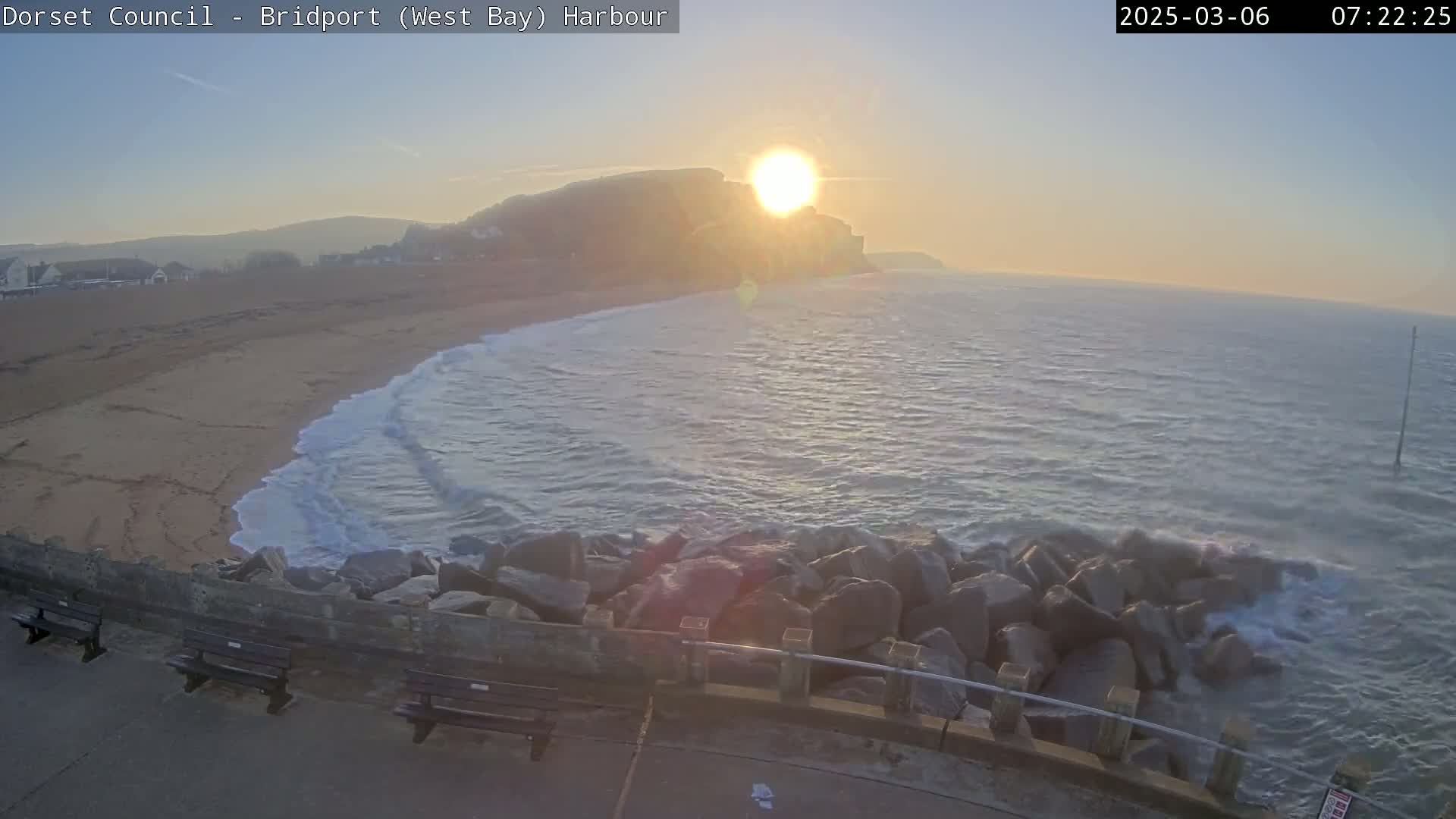 A sunrise illuminates a sandy beach and calm ocean waves breaking against a rocky breakwater, with a cliff face visible in the distance.
