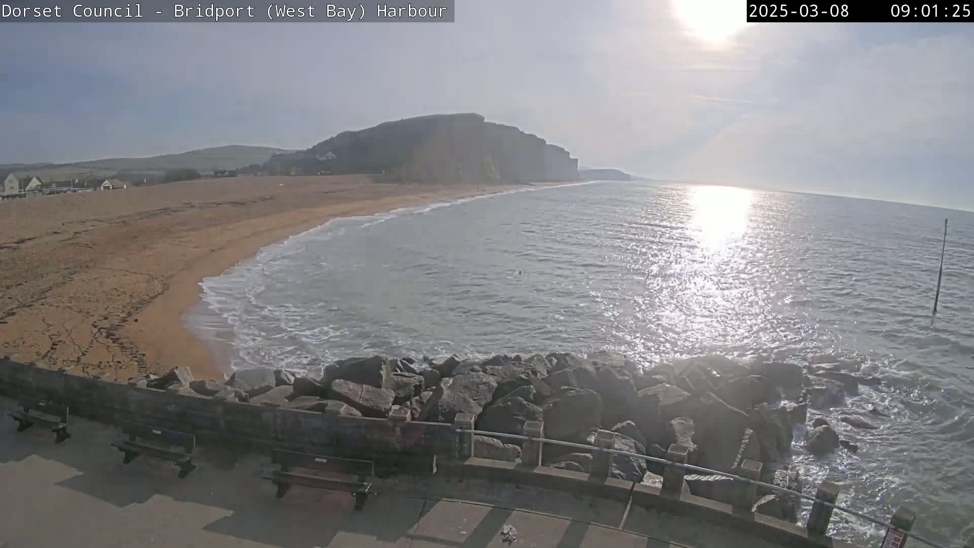 A sandy beach meets a wavy sea under a bright, sunny sky, with a rocky seawall and cliffs in the background.