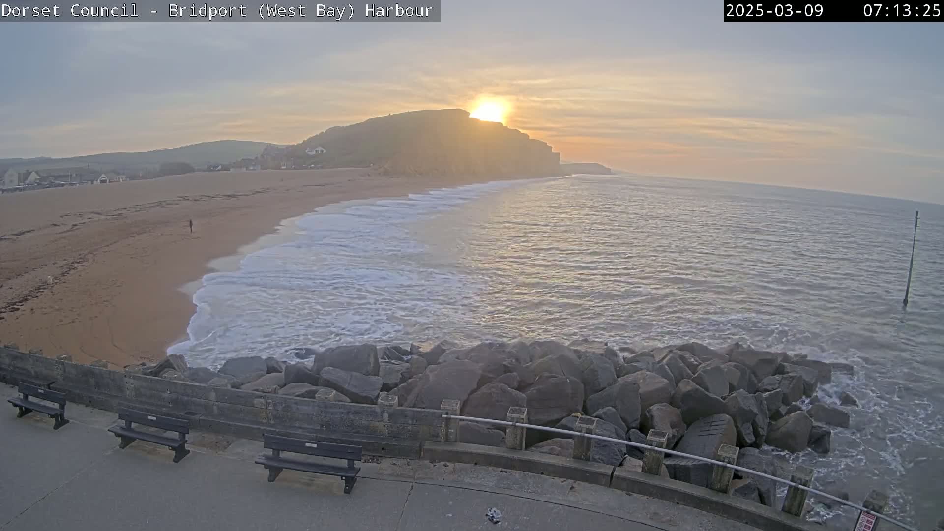 A sandy beach with gentle waves meets a rocky seawall under a sunrise sky.