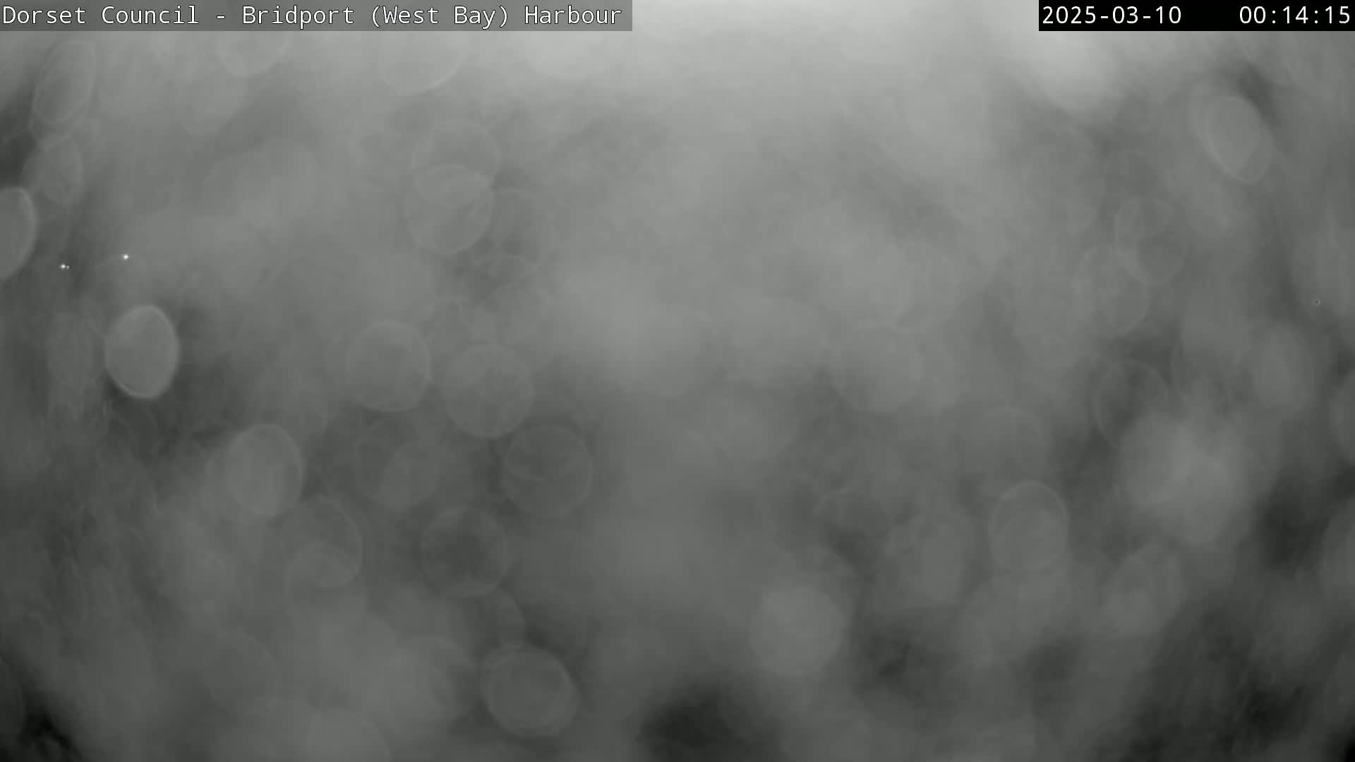 A blurry, out-of-focus image shows a dark gray scene with many indistinct light-gray circles, likely raindrops on a lens, suggesting rainy weather conditions.