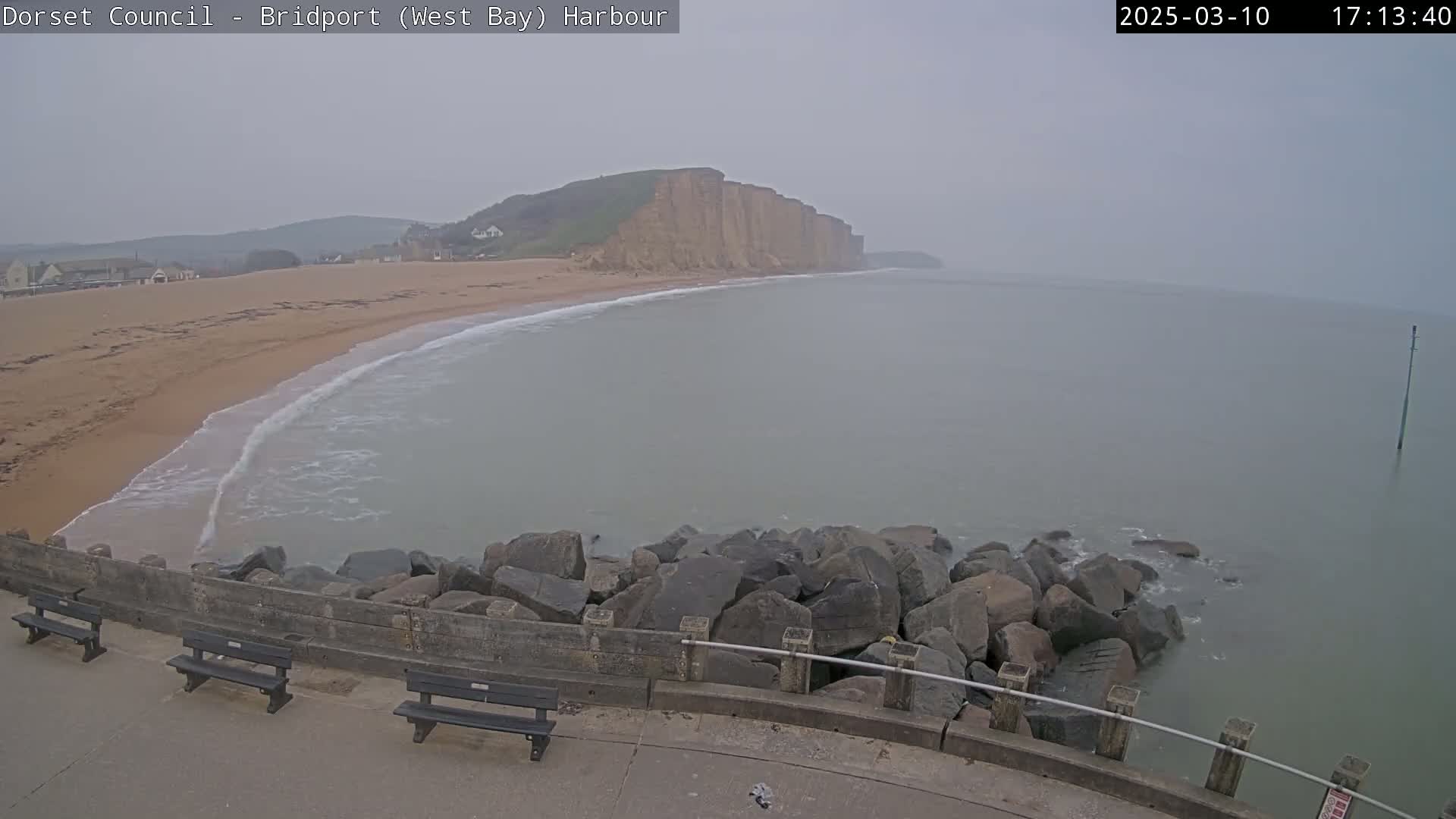 A hazy, overcast day reveals a sandy beach curving towards tall, pale cliffs overlooking a calm sea, with a rocky breakwater and several park benches in the foreground.