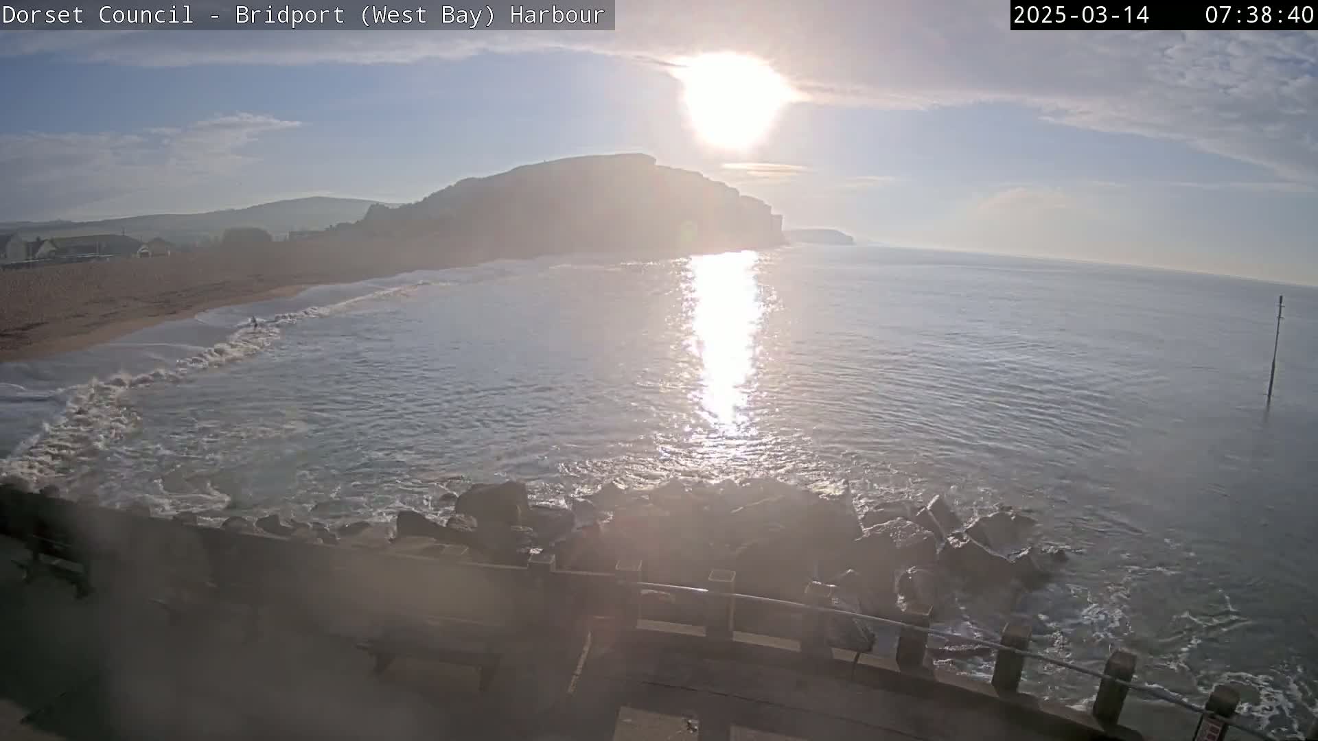 A bright, sunny morning at a harbor shows calm ocean waves lapping against a rocky shore with a sandy beach and distant cliffs visible.