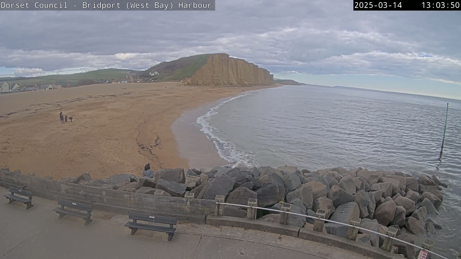 A sandy beach curves gently into a calm, gray sea under a cloudy sky, backed by a tall, cliff face.