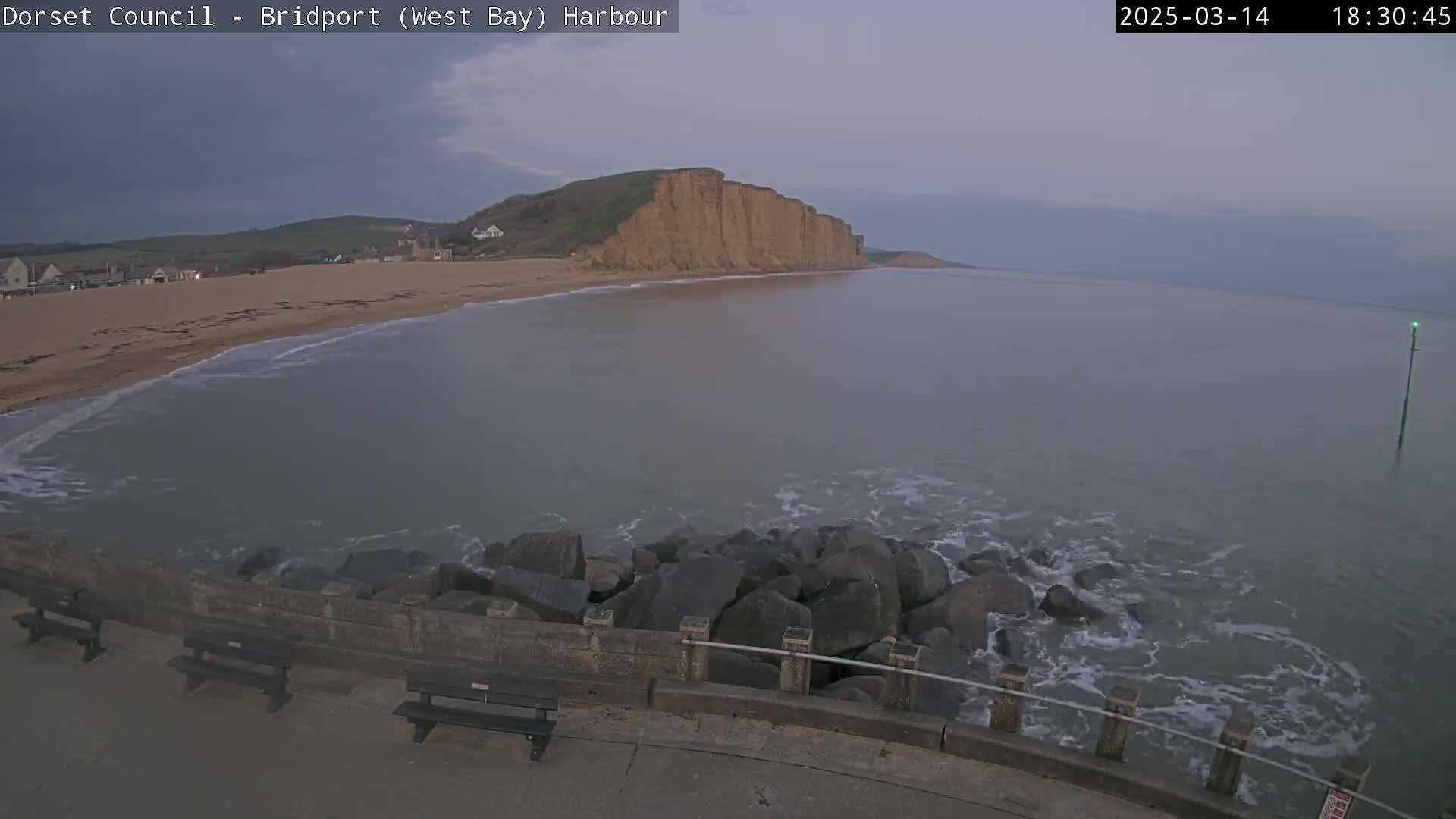 A calm, overcast evening scene shows a sandy beach, a rocky seawall, and tall cliffs overlooking a bay with gentle waves.