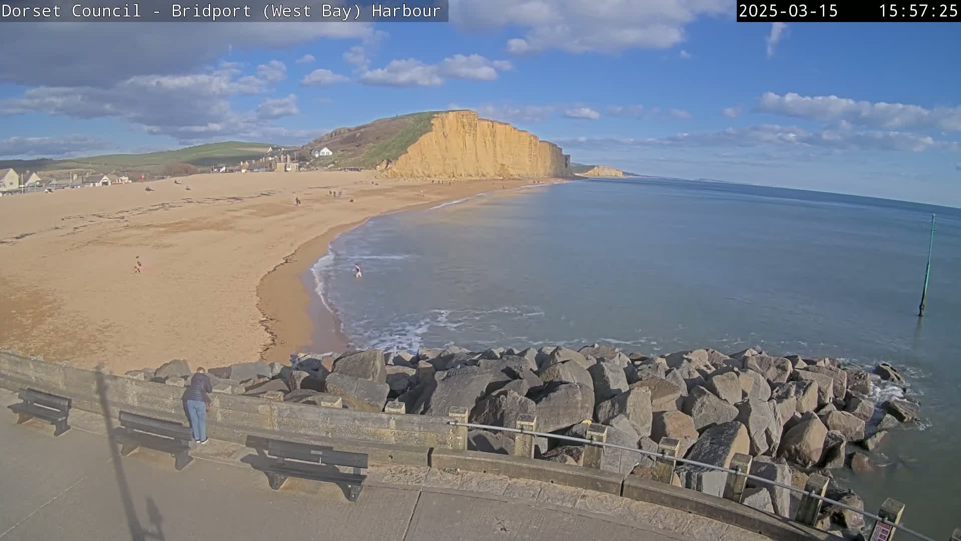 A sandy beach stretches beneath a partly cloudy blue sky, meeting a calm sea protected by a rock barrier, with a large cliff face visible in the distance.