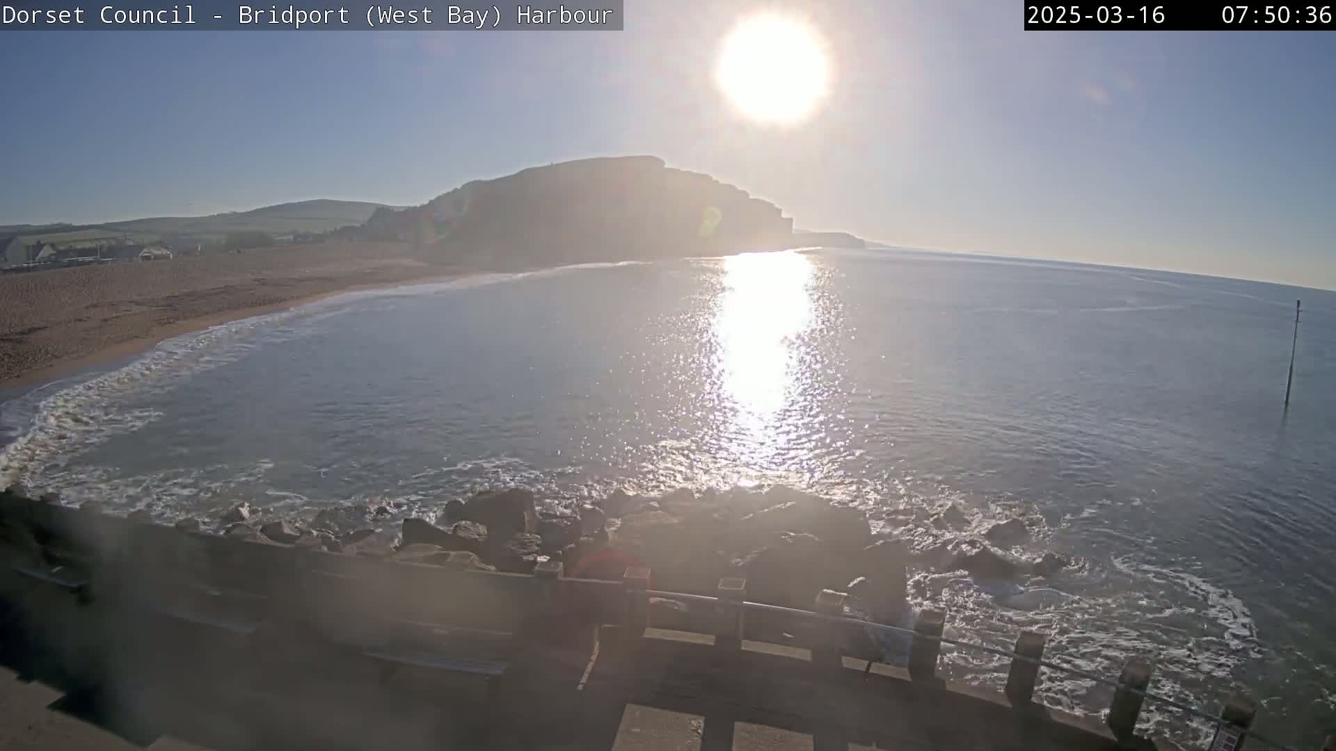 A bright, sunny day reveals a calm bay with a sandy beach and cliffs in the background, viewed from a rocky harbor wall.