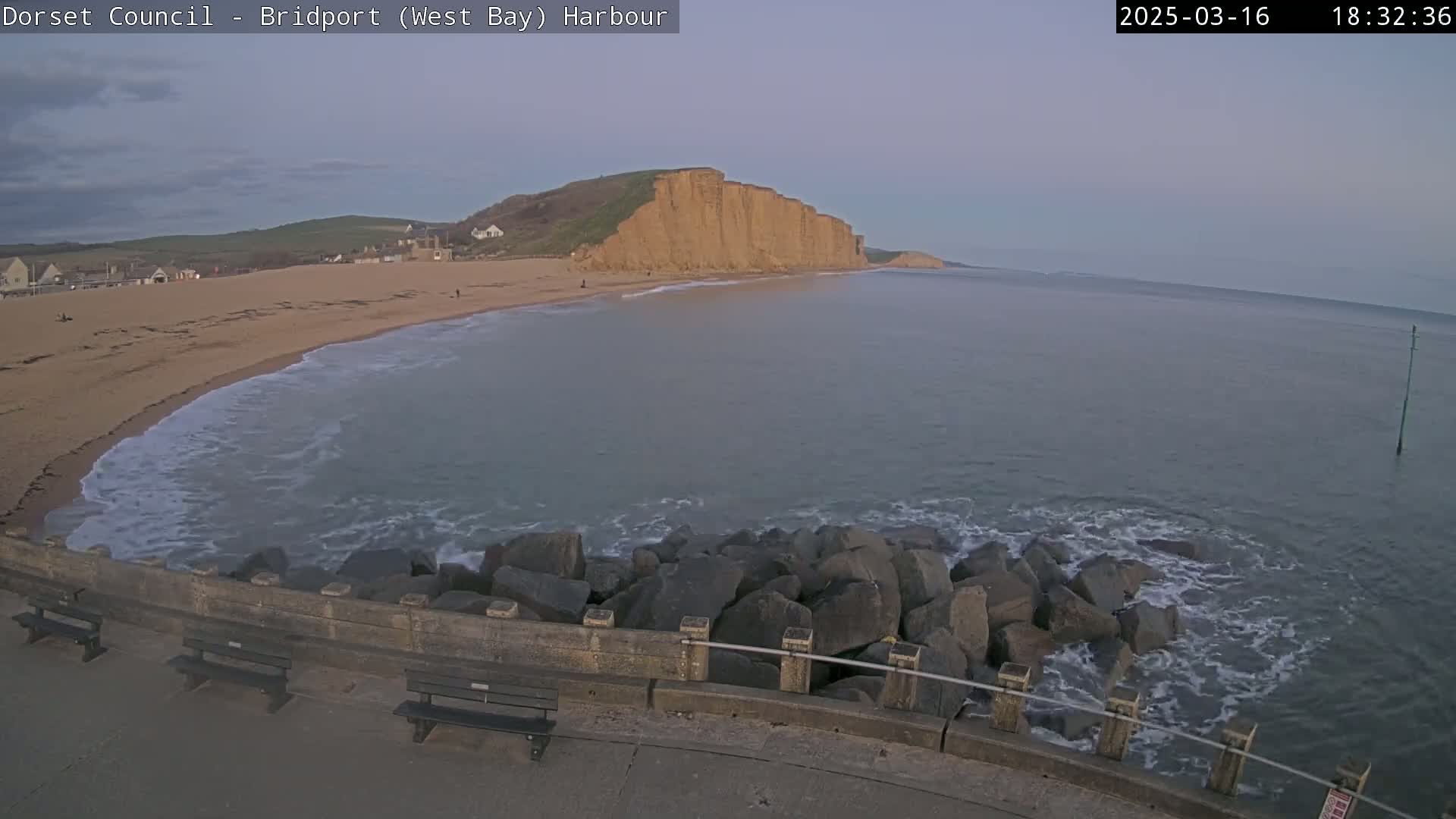 A sandy beach curves gently into a calm sea under a dusky sky, with a large cliff face visible in the distance and a rock breakwater in the foreground.