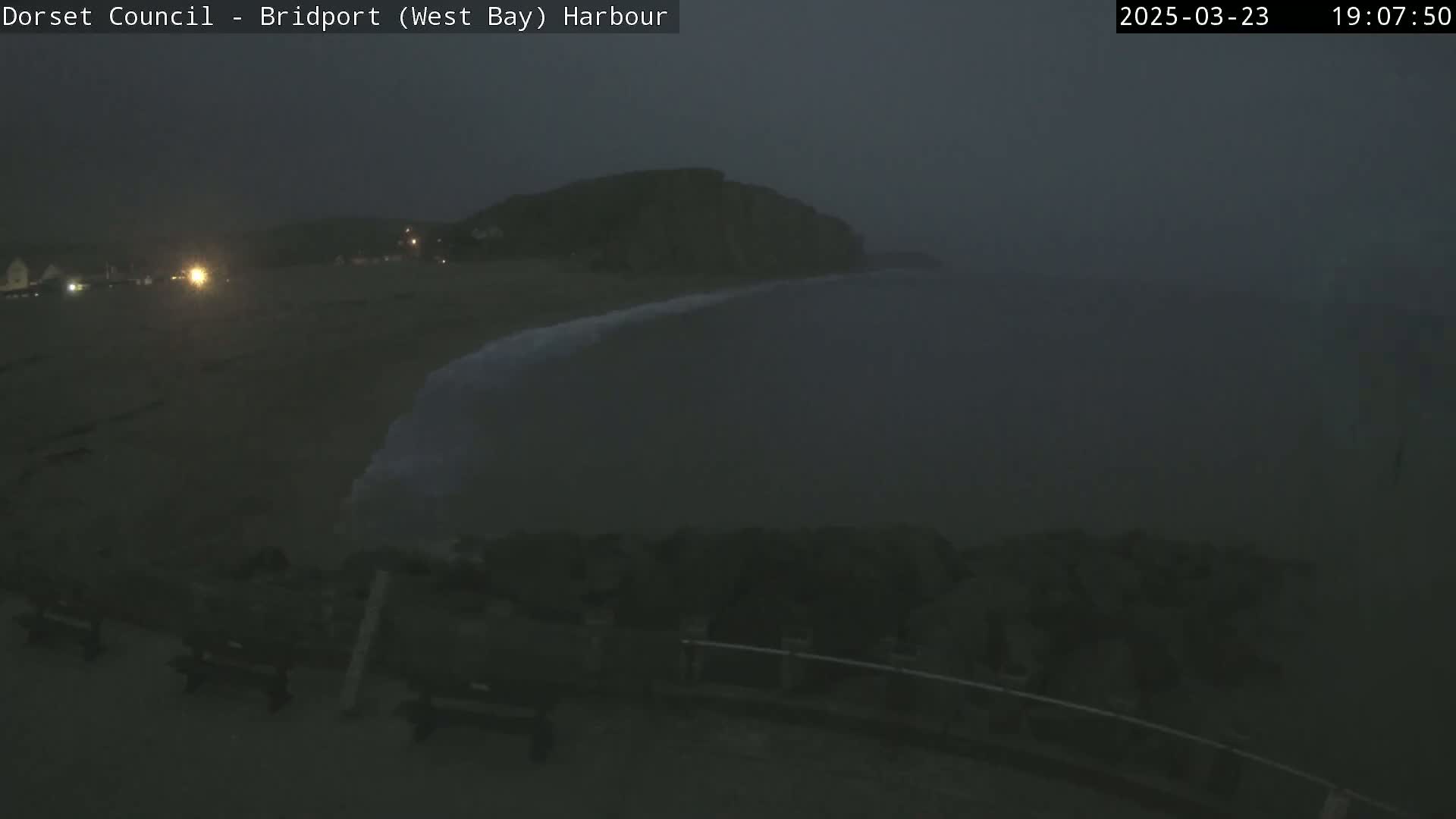 A dark, foggy night scene shows a beach with a low cliff in the distance and a few benches in the foreground.
