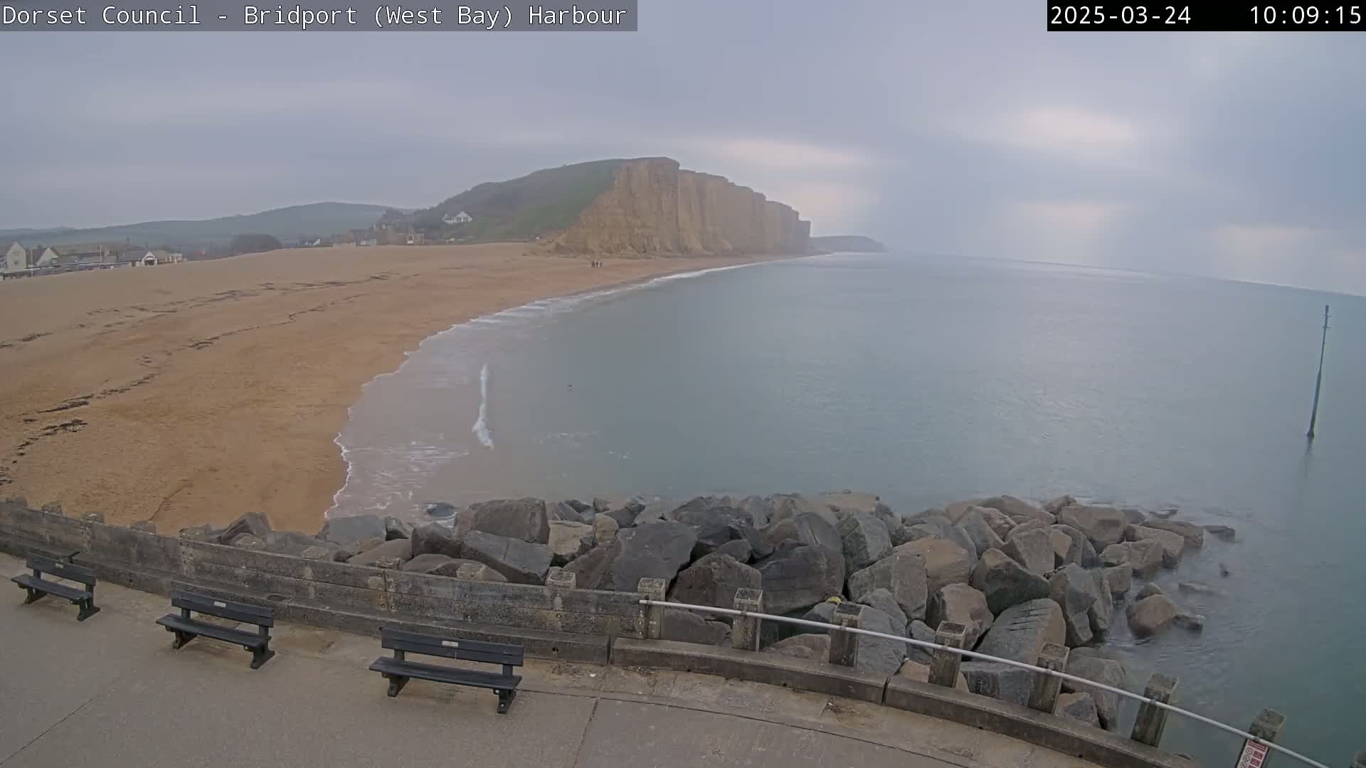 A sandy beach curves around a calm, gray sea under an overcast sky, with a high cliff visible in the distance and large rocks forming a breakwater in the foreground.