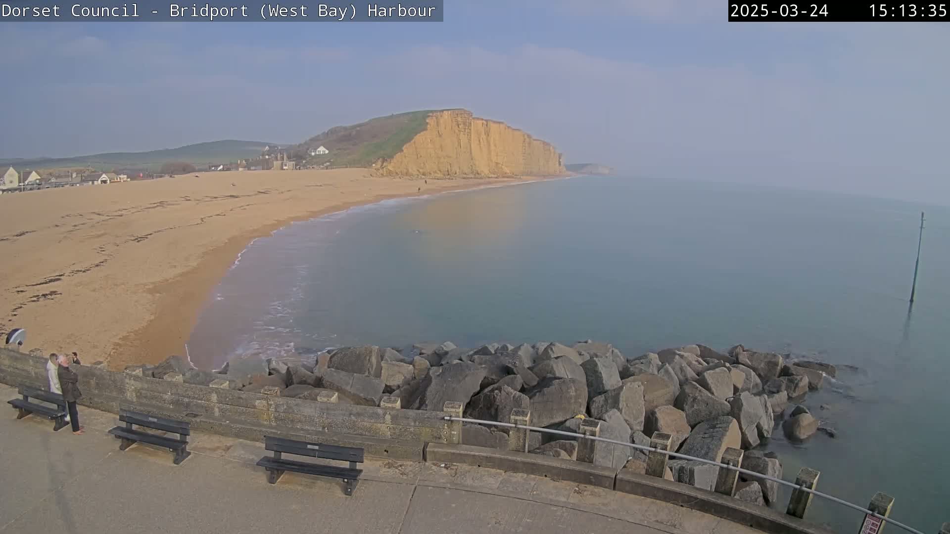 A sandy beach stretches to a calm, hazy ocean under a mostly clear sky, backed by a steep, tan cliff.