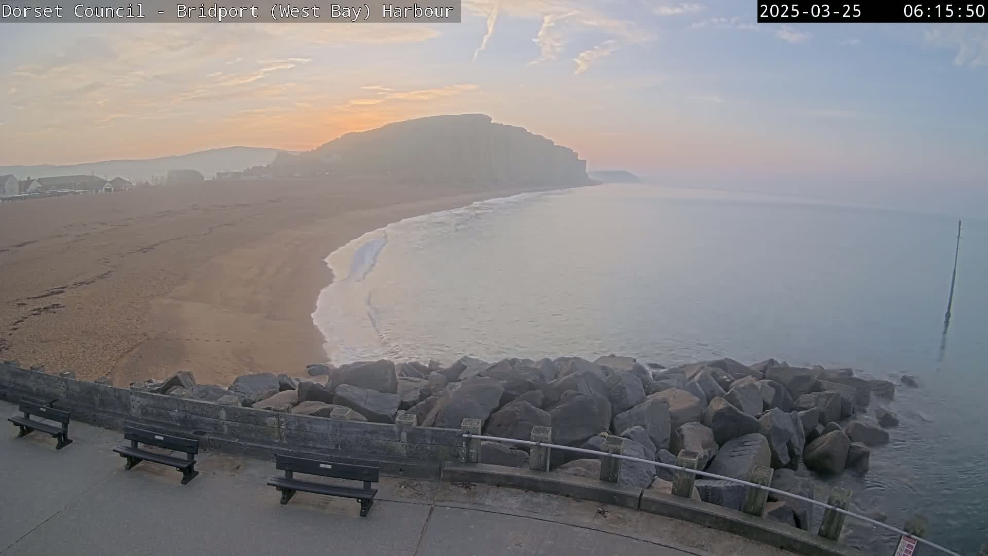 A sandy beach curves around a calm, hazy sea towards a large cliff face at sunrise.