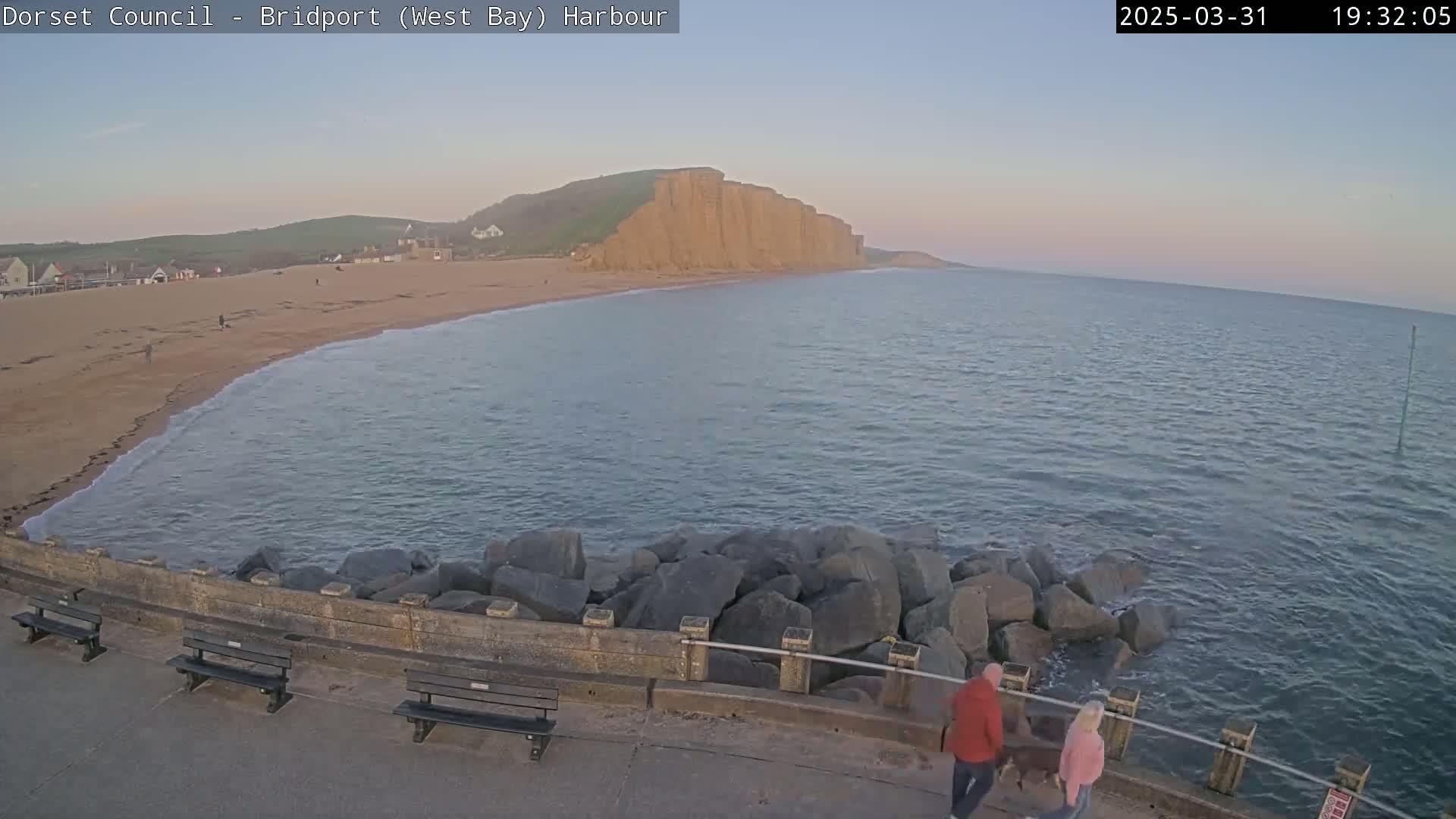 A sandy beach curves gently around a calm, twilight sea, backed by tall, reddish-brown cliffs.