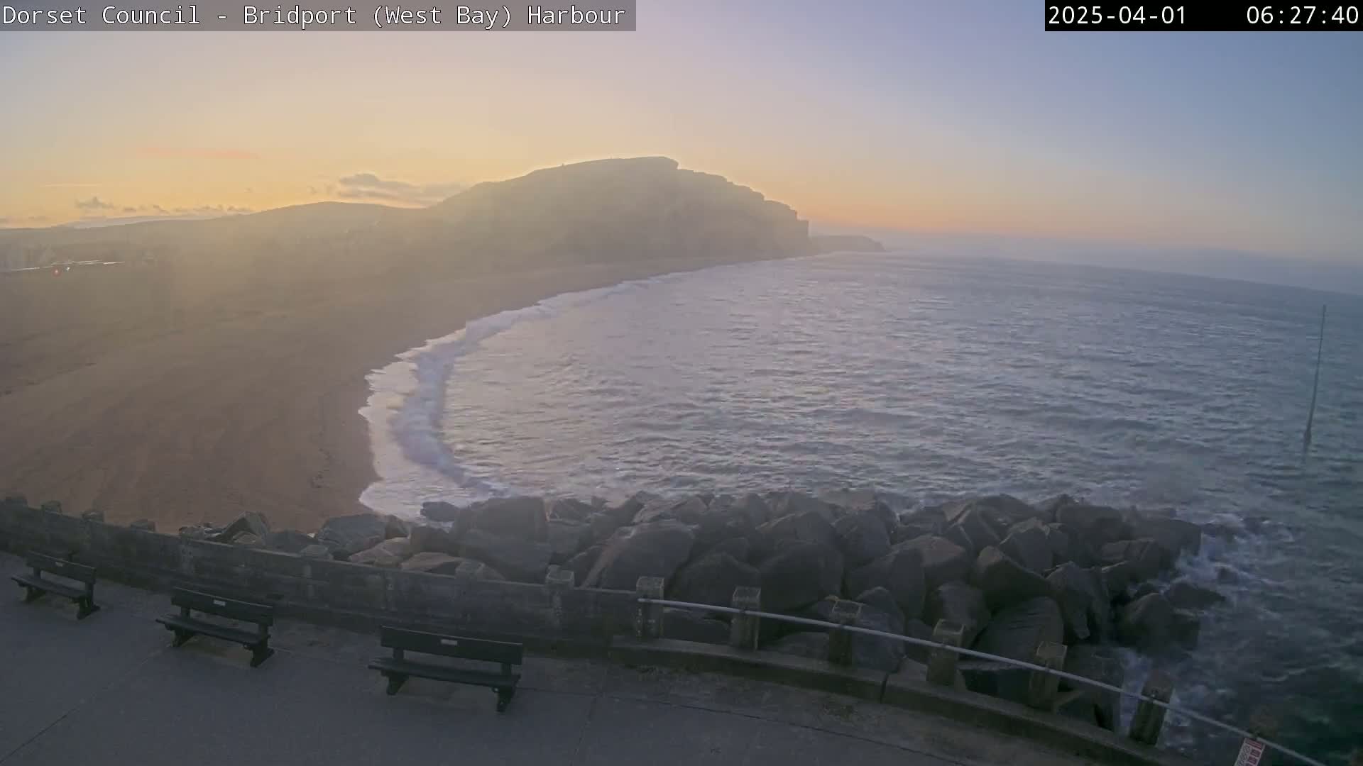 A hazy sunrise illuminates a sandy beach, gentle waves lapping against a rocky breakwater, with cliffs visible in the distance.