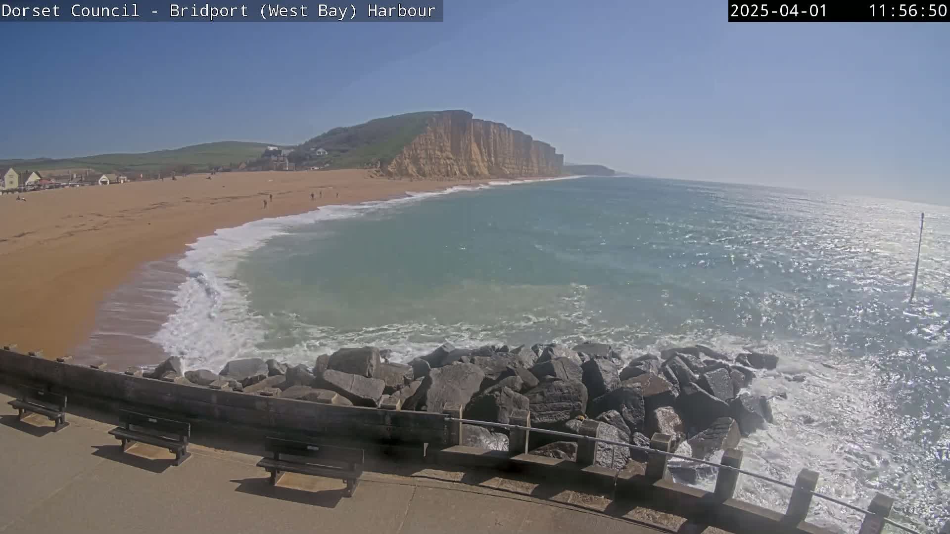 A sandy beach with gentle waves laps against a rocky breakwater under a clear blue sky.