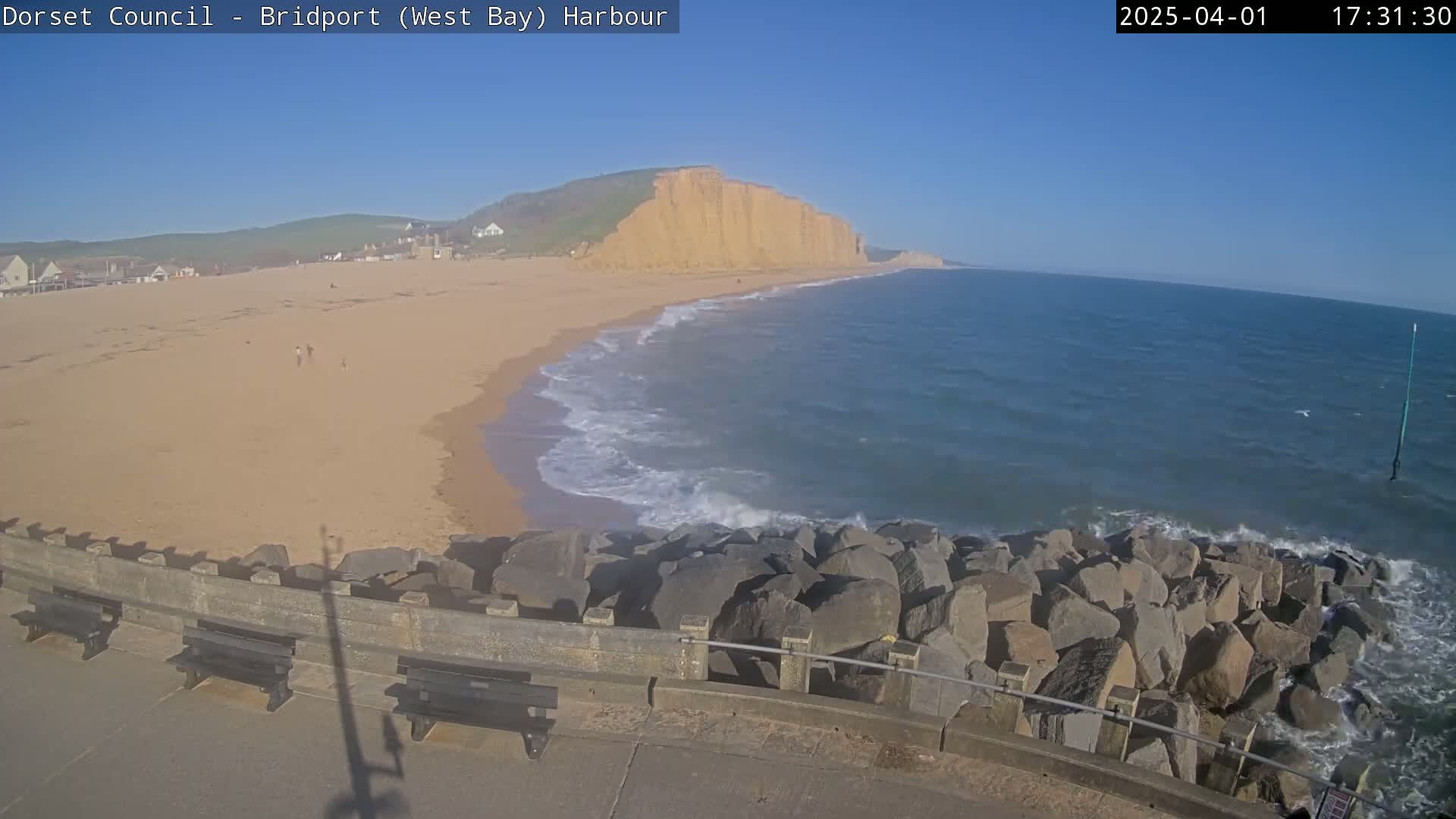 A sandy beach with a few people on it stretches beneath a large cliff face, bordered by a rocky breakwater where small waves are lapping against it under a clear blue sky.