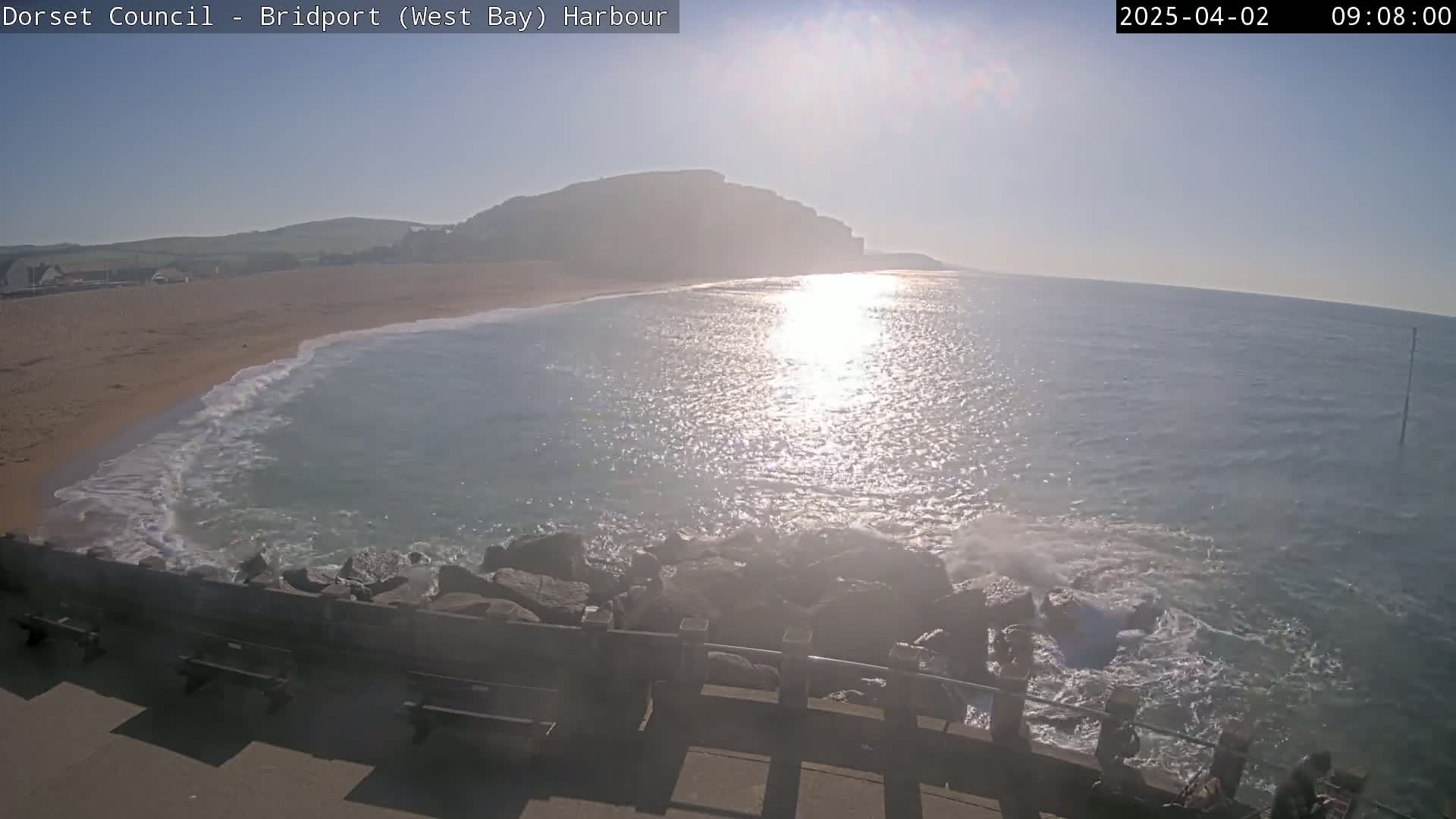 A sunny day at a harbor shows a sandy beach with gentle waves lapping against rocks, a calm sea reflecting sunlight, and a large hill in the background.