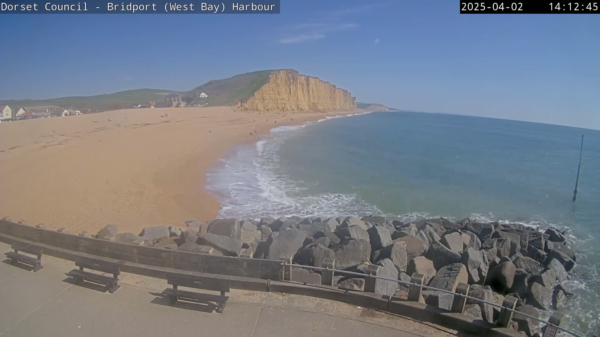 A sandy beach stretches out to meet a calm ocean under a clear blue sky, with large rocks forming a barrier between the beach and a paved walkway lined with benches.
