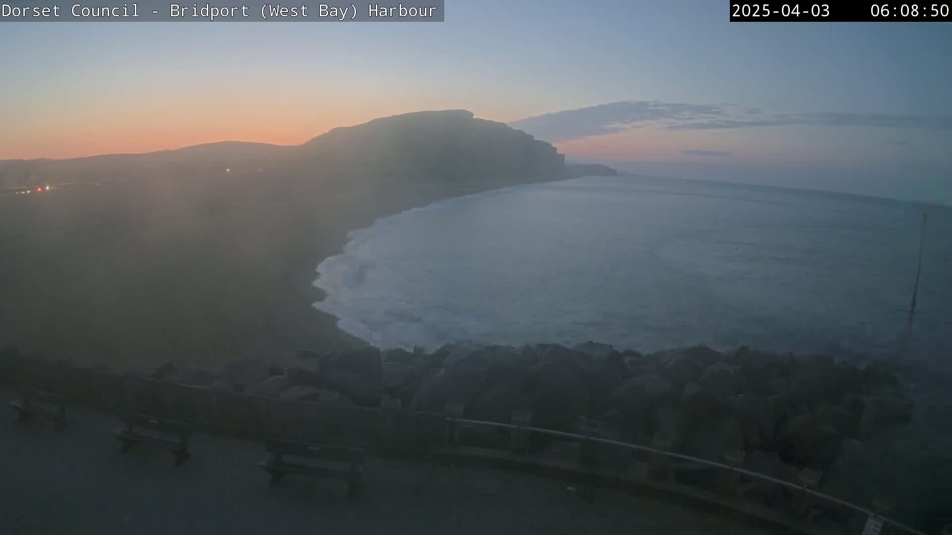 A hazy sunrise illuminates a calm bay with gentle waves breaking against a rocky shore, where several benches sit beside a low wall.