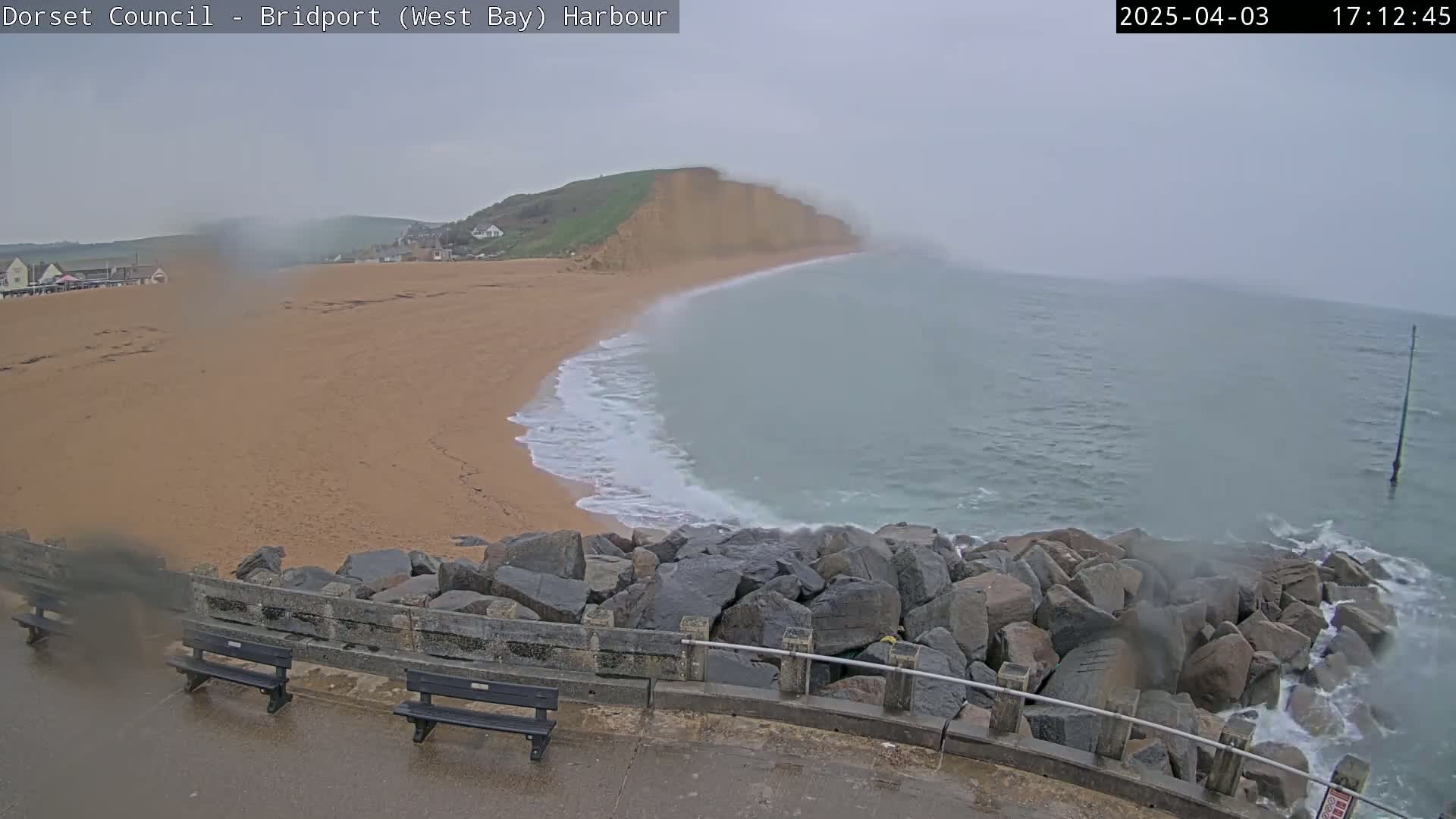 A sandy beach curves around a rocky breakwater into a grey, overcast ocean meeting a large cliff face.