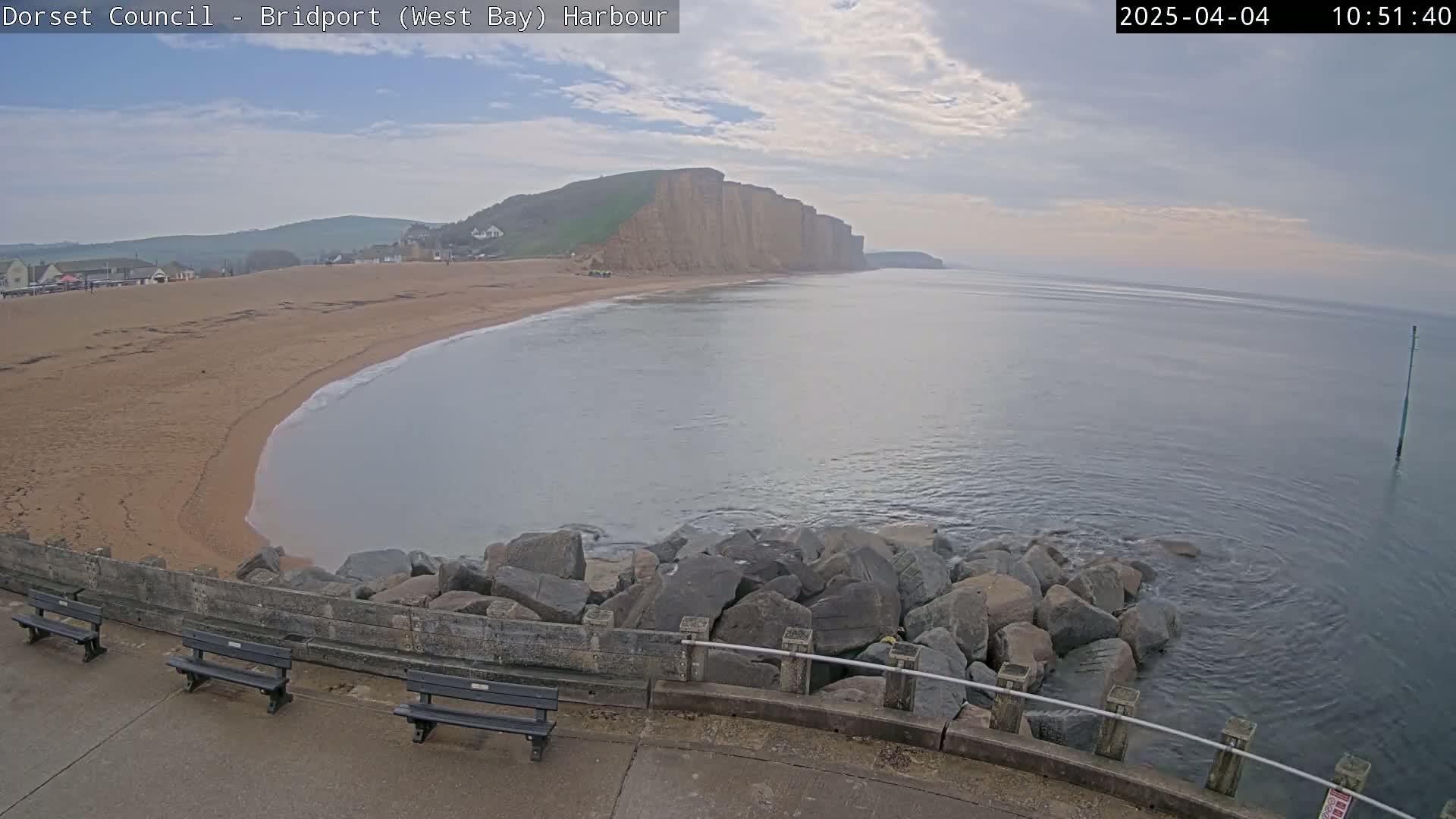 A sandy beach curves into a calm bay, bordered by a rocky breakwater and overlooking imposing cliffs under a partly cloudy sky.