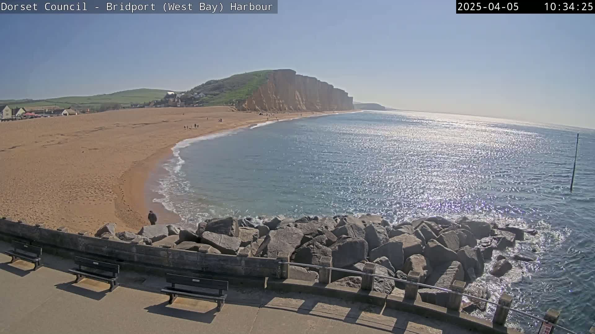 A sandy beach stretches alongside a calm, sunlit ocean, backed by a high, grassy cliff face on a sunny day.