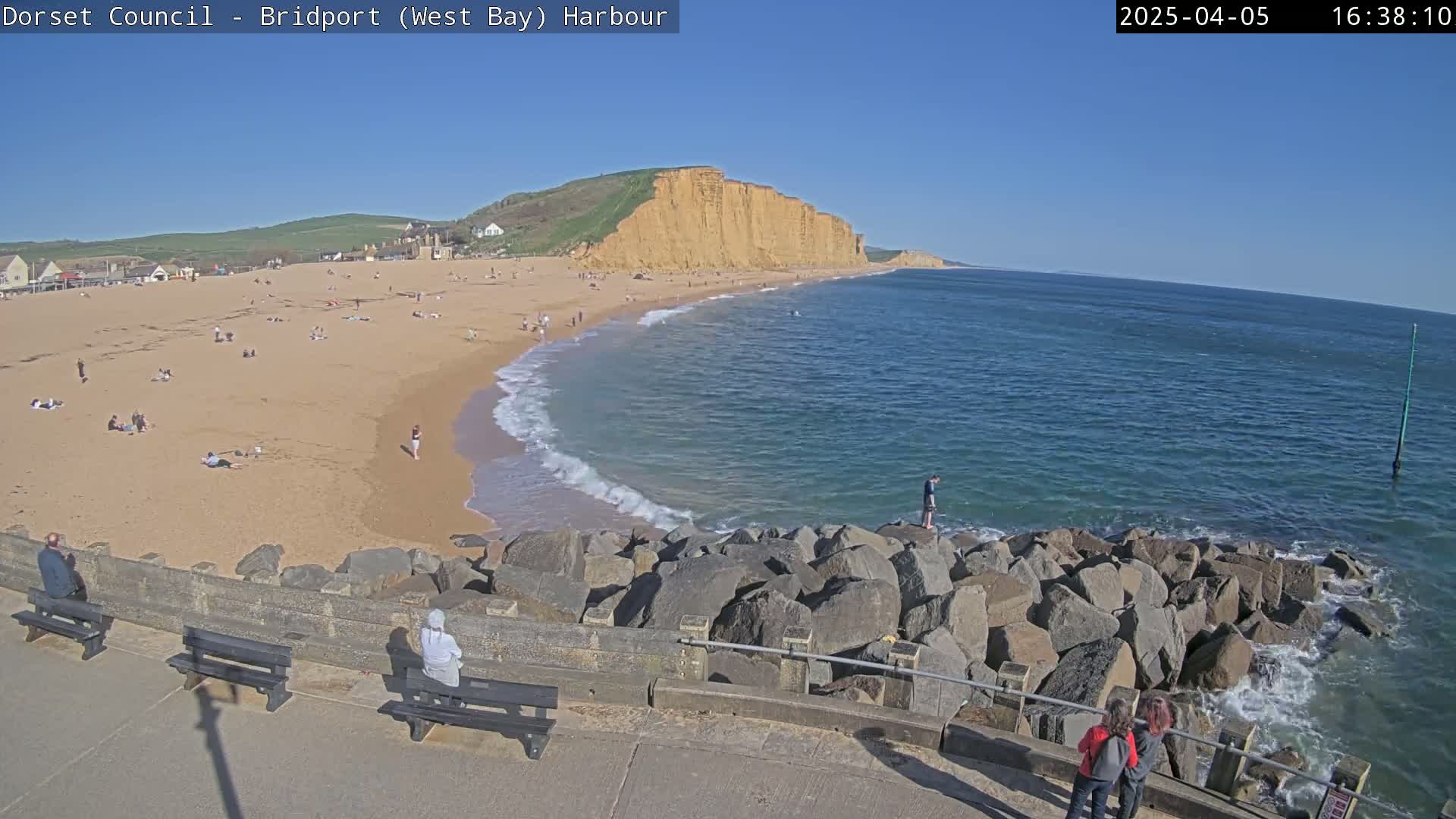 A sandy beach with people sunbathing and swimming, backed by cliffs, under a clear blue sky.