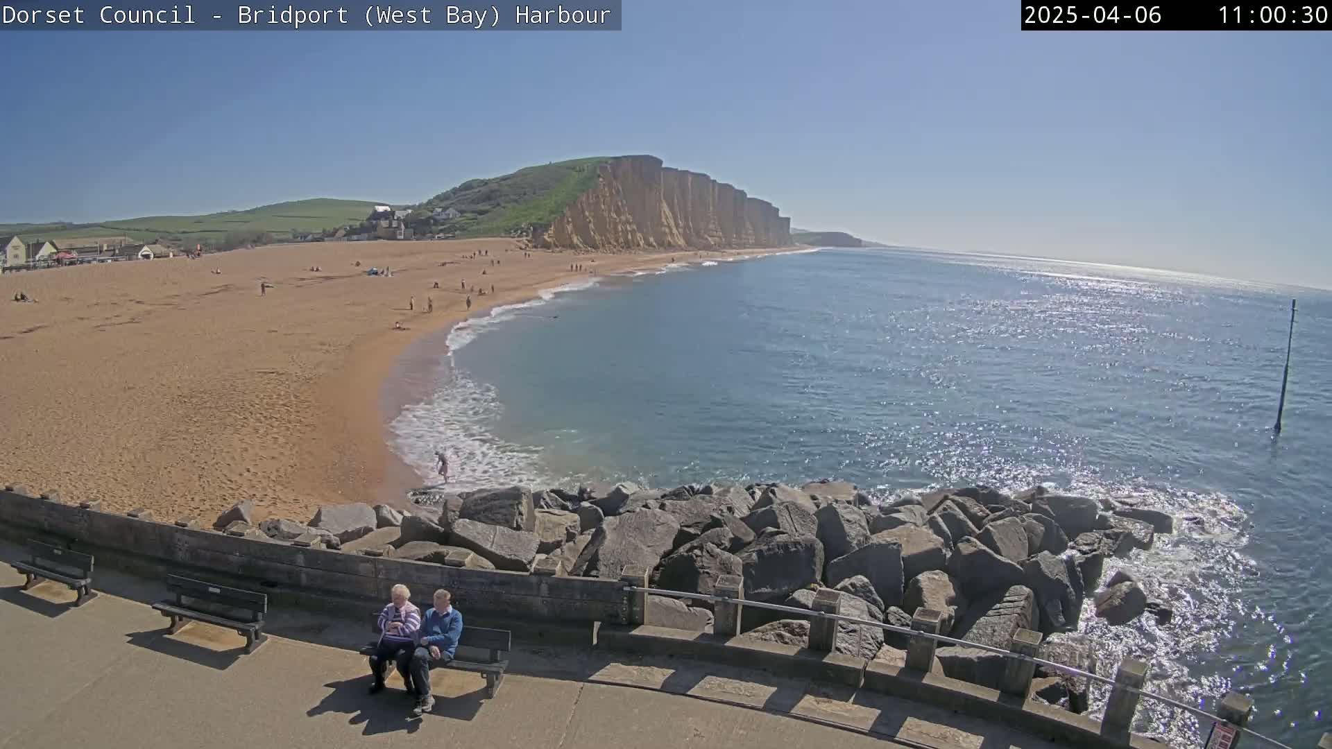 A sunny day at a sandy beach with a rocky shoreline and cliffs in the background, featuring a couple sitting on a bench.
