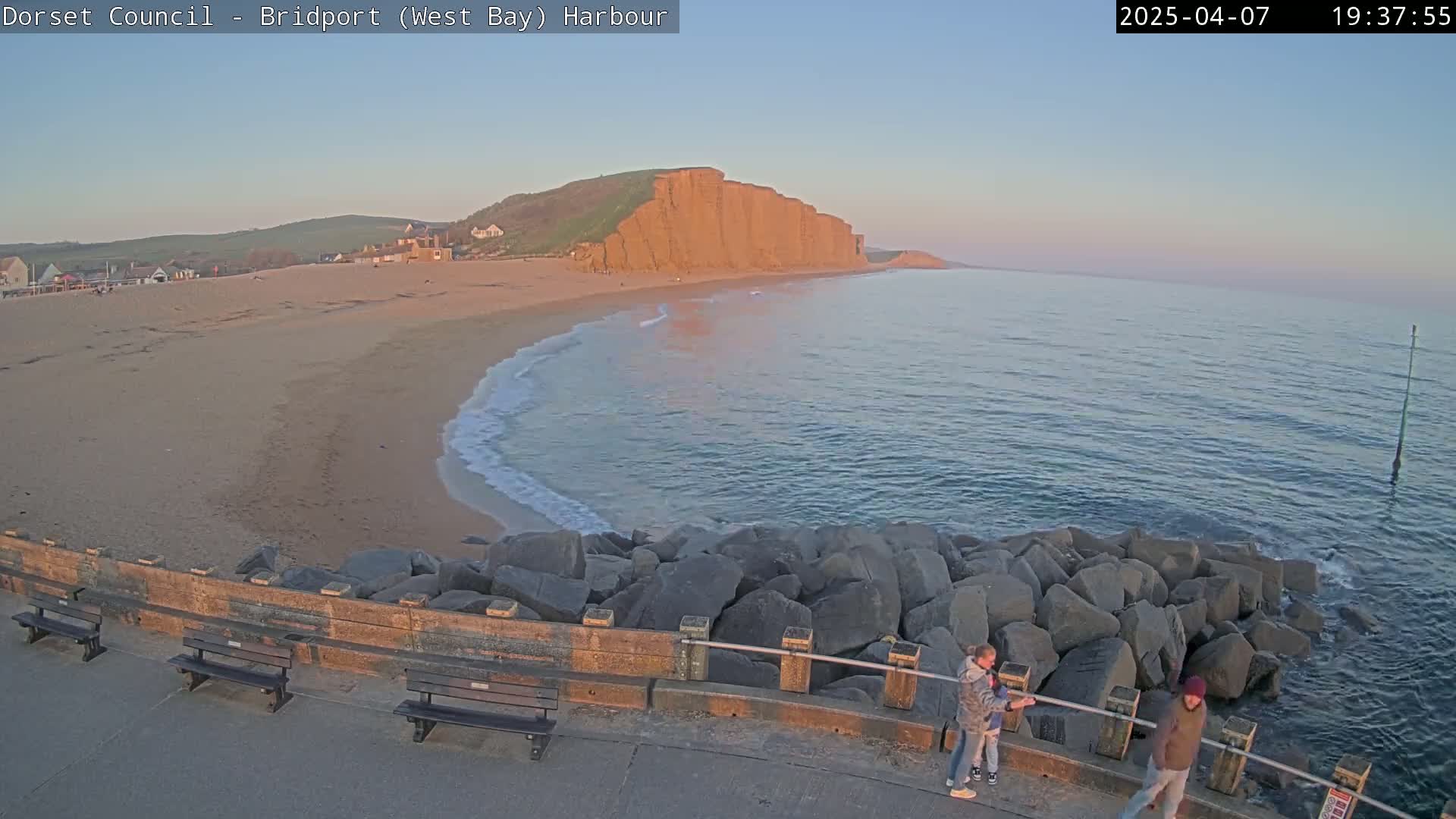 A sandy beach curves around a rocky breakwater, with a large, reddish cliff in the background, under a clear, twilight sky.