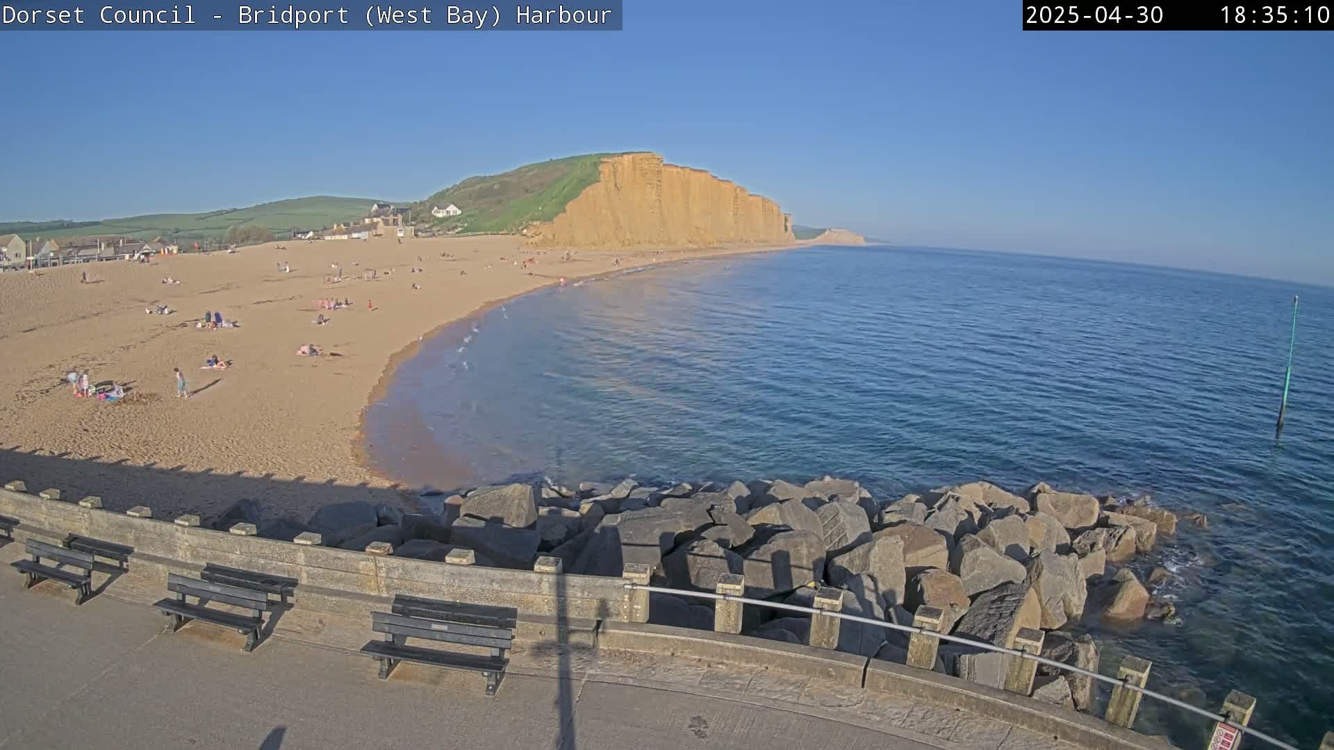 A sandy beach with people on it, backed by cliffs, meets a calm blue sea under a clear blue sky.